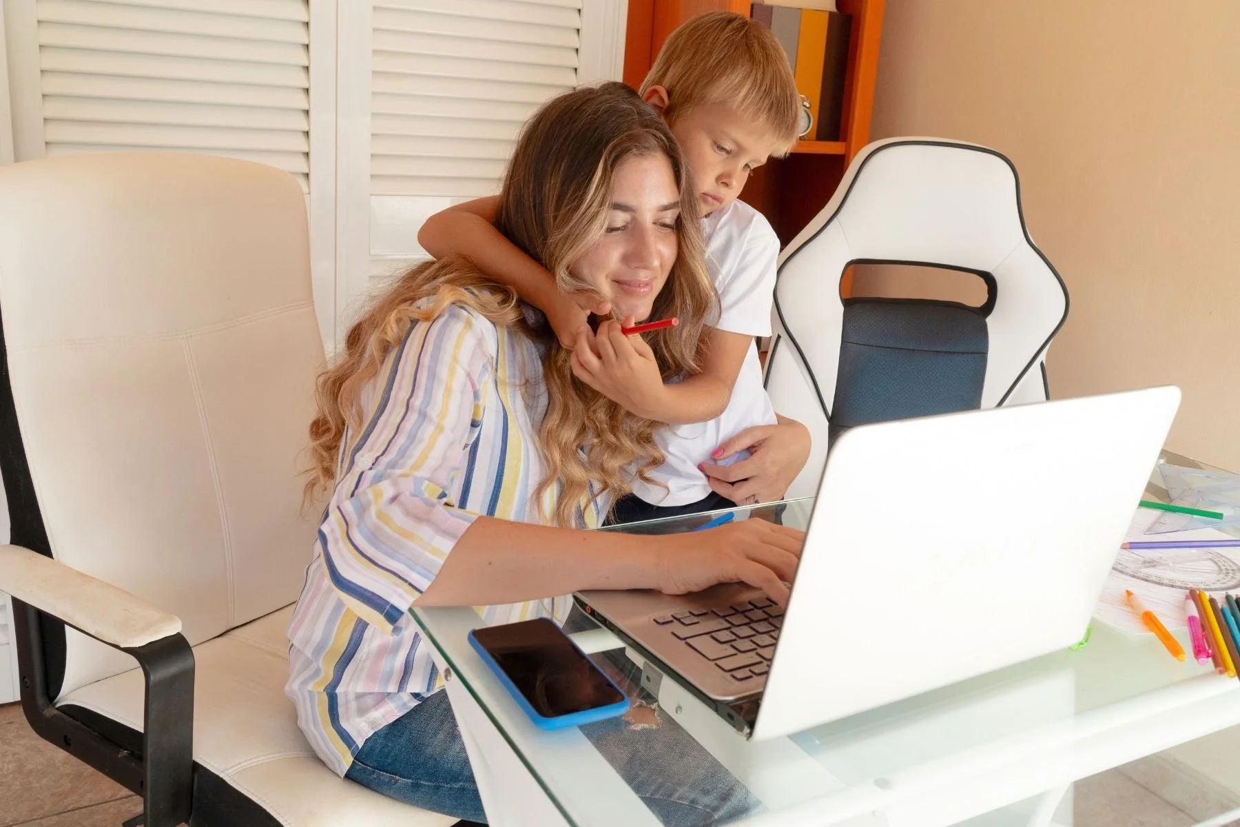 A woman and a young boy sitting at a glass desk, looking at a laptop screen. The woman is smiling slightly, and the boy is hugging her from behind, holding a red pen. The desk has various colorful pens, papers, a phone, and a laptop. The setting appears to be a home office or study area.