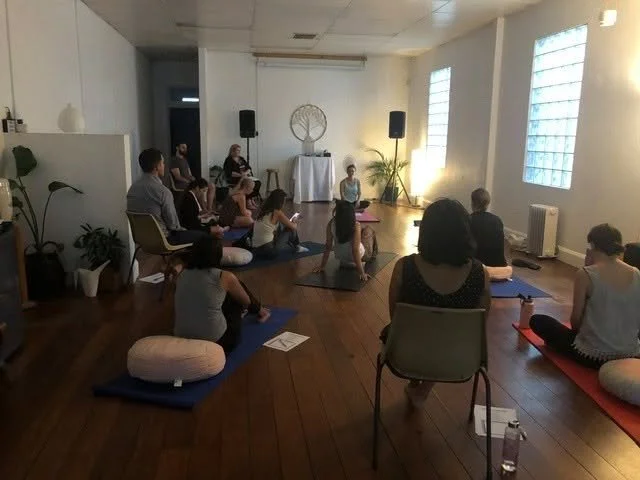 Group of people participating in a yoga or meditation class in a bright room with wooden flooring, large windows, and minimal decor.