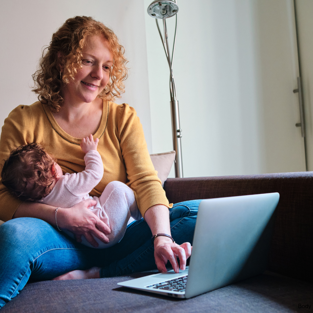A woman with curly red hair sitting on a couch, holding a small baby, while looking at a laptop screen.