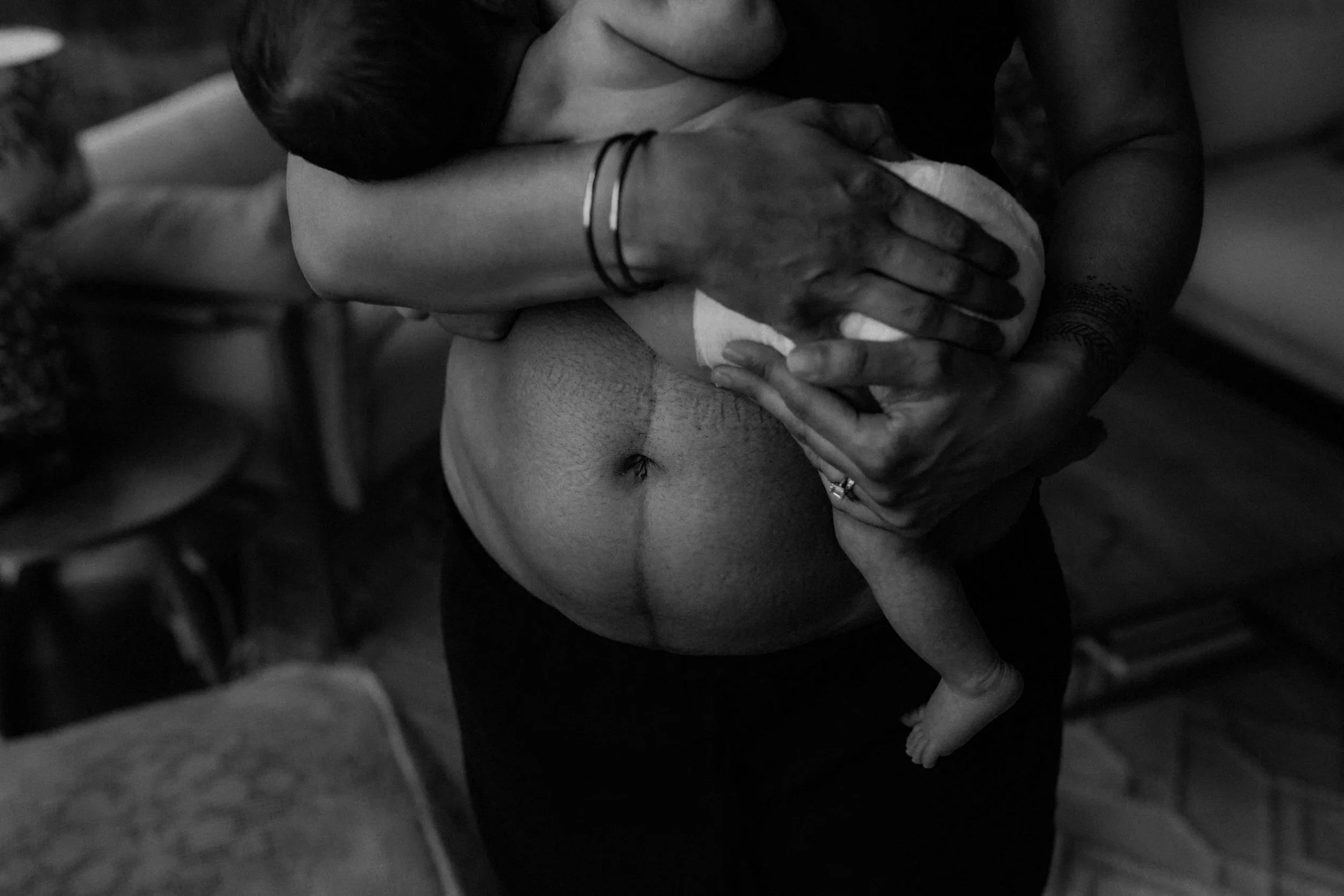 A person holding a small baby, with the baby's head resting on their shoulder and the baby's body dangling downward, showing the back and legs of the baby in black and white.