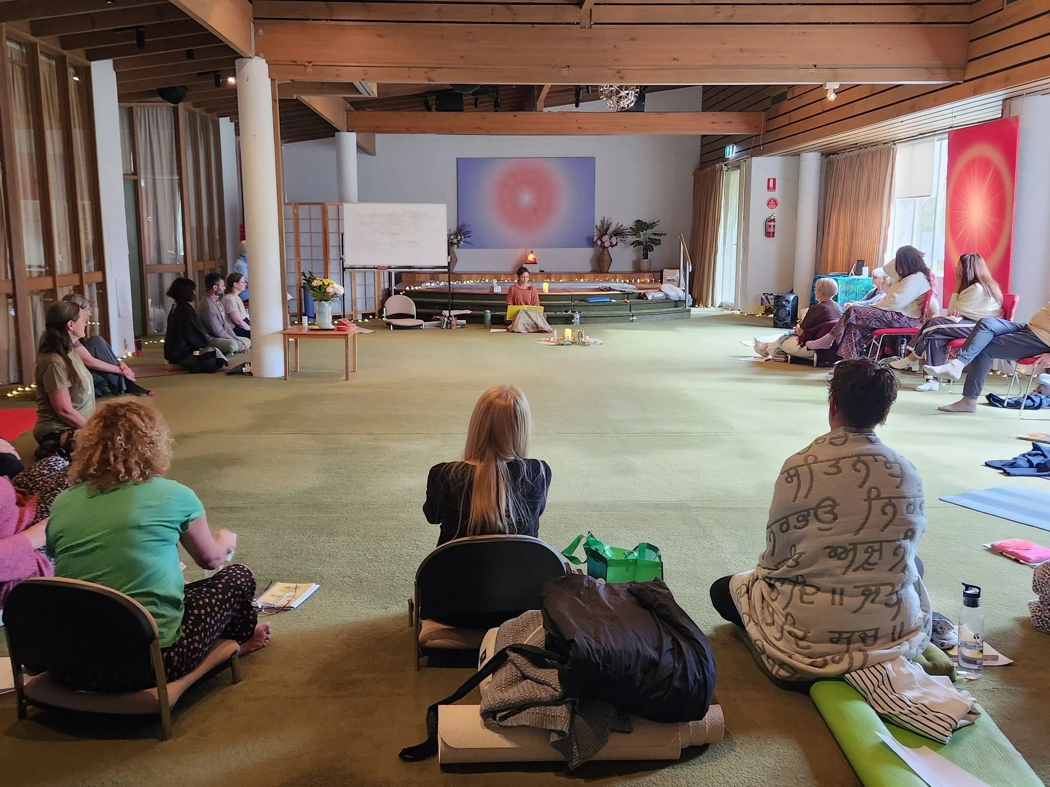 People seated on the floor and chairs in a room attending a workshop or class, with a woman at the front, a whiteboard, and colorful artwork on the wall.
