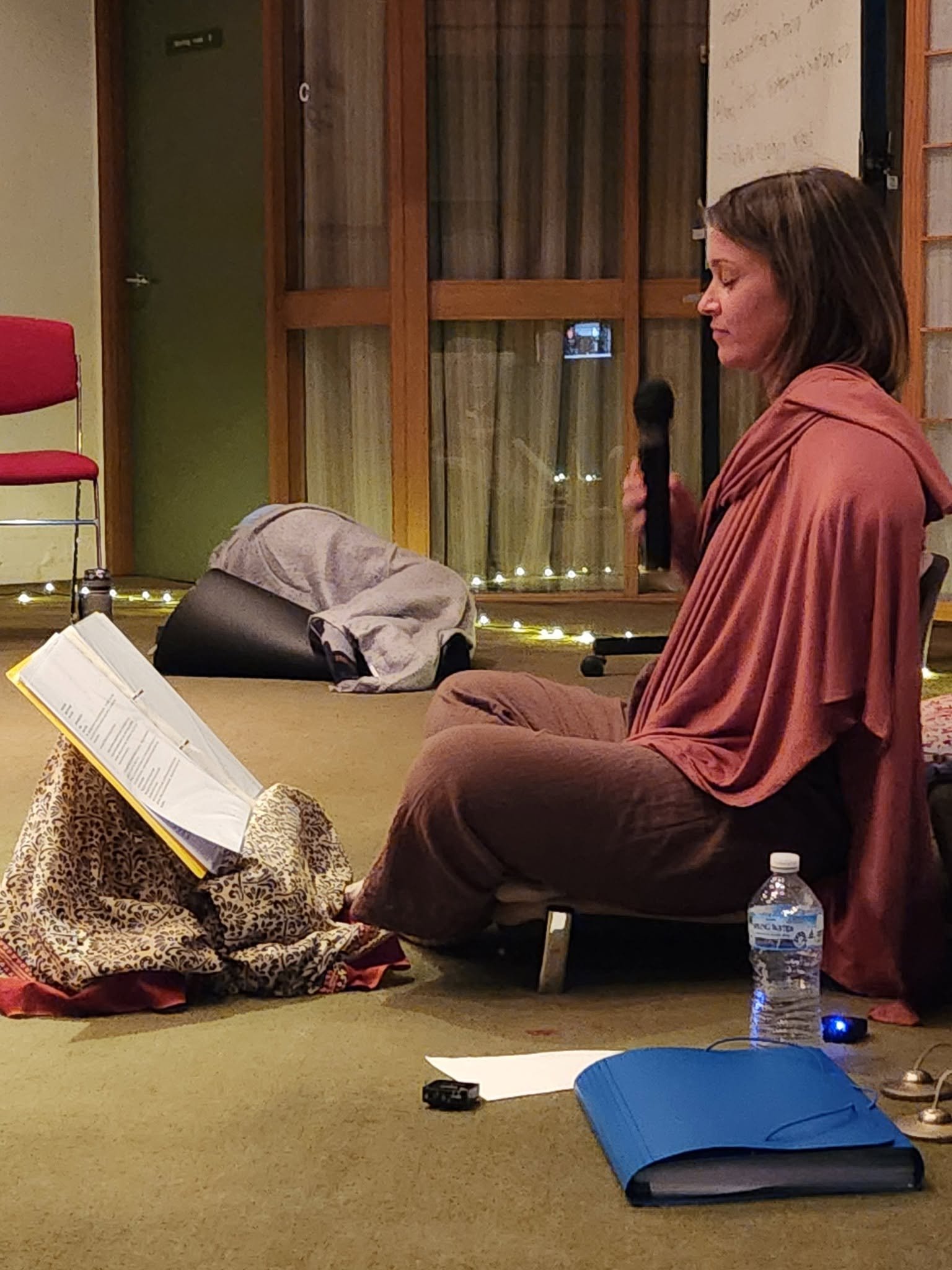 Woman sitting cross-legged on the floor with eyes closed, holding a microphone, near a whiteboard and window with curtains and string lights, with a water bottle and blue folder nearby.