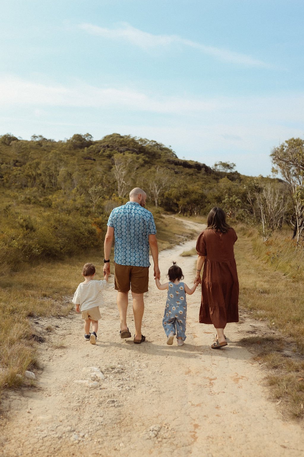 A family of four walking on a dirt trail through a natural landscape with trees and hills, under a blue sky.
