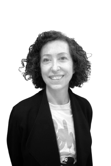 A smiling woman with her arms down by her sides. She has cropped, curly hair, and is wearing a blazer and t-shirt with a squiggly print on it. The image is cropped to waist height and is in greyscale.