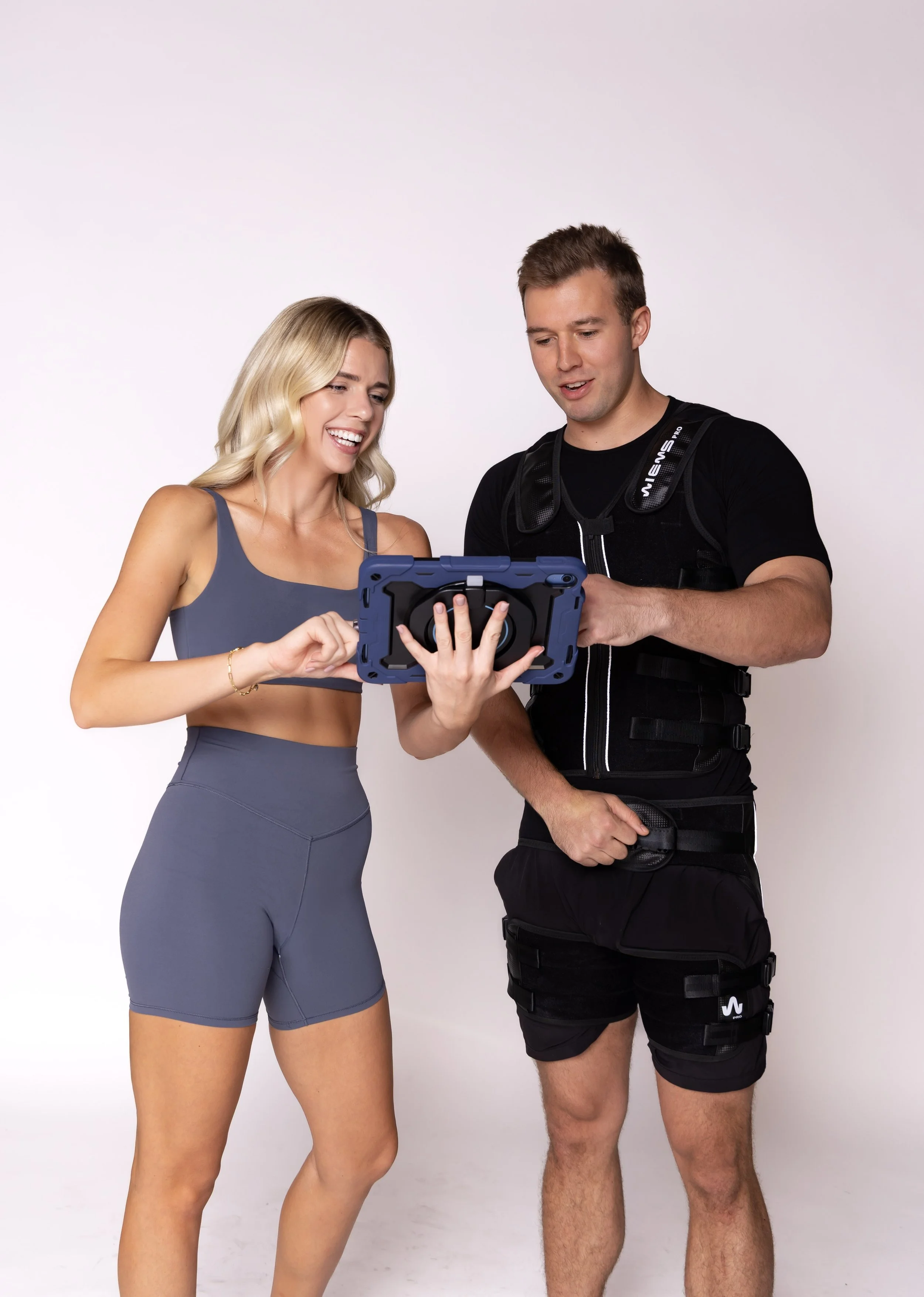 A woman and a man are looking at a tablet together smiling, in workout attire, against a plain white background.
