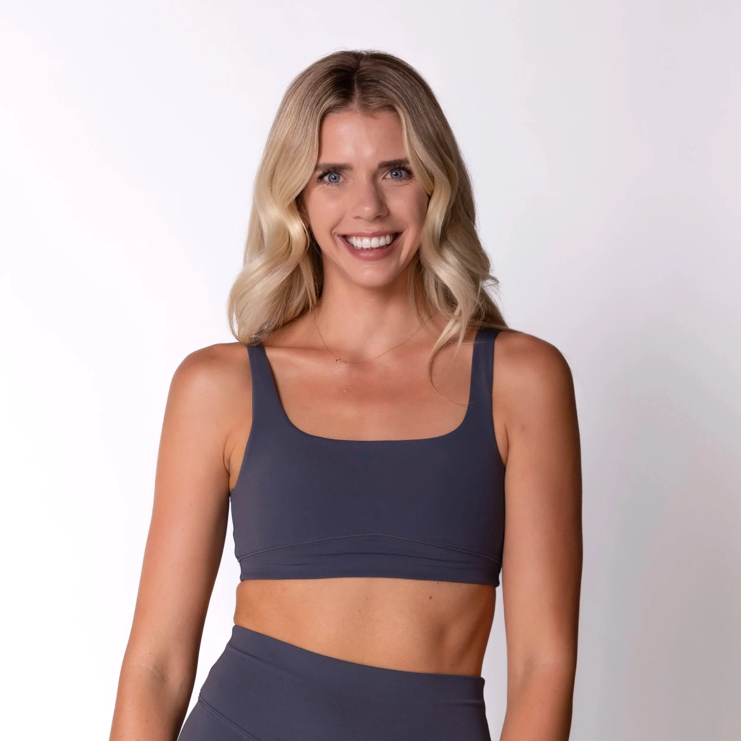 A young blonde woman with wavy hair smiling, wearing a white sports crop top and black pants, against a plain white background.