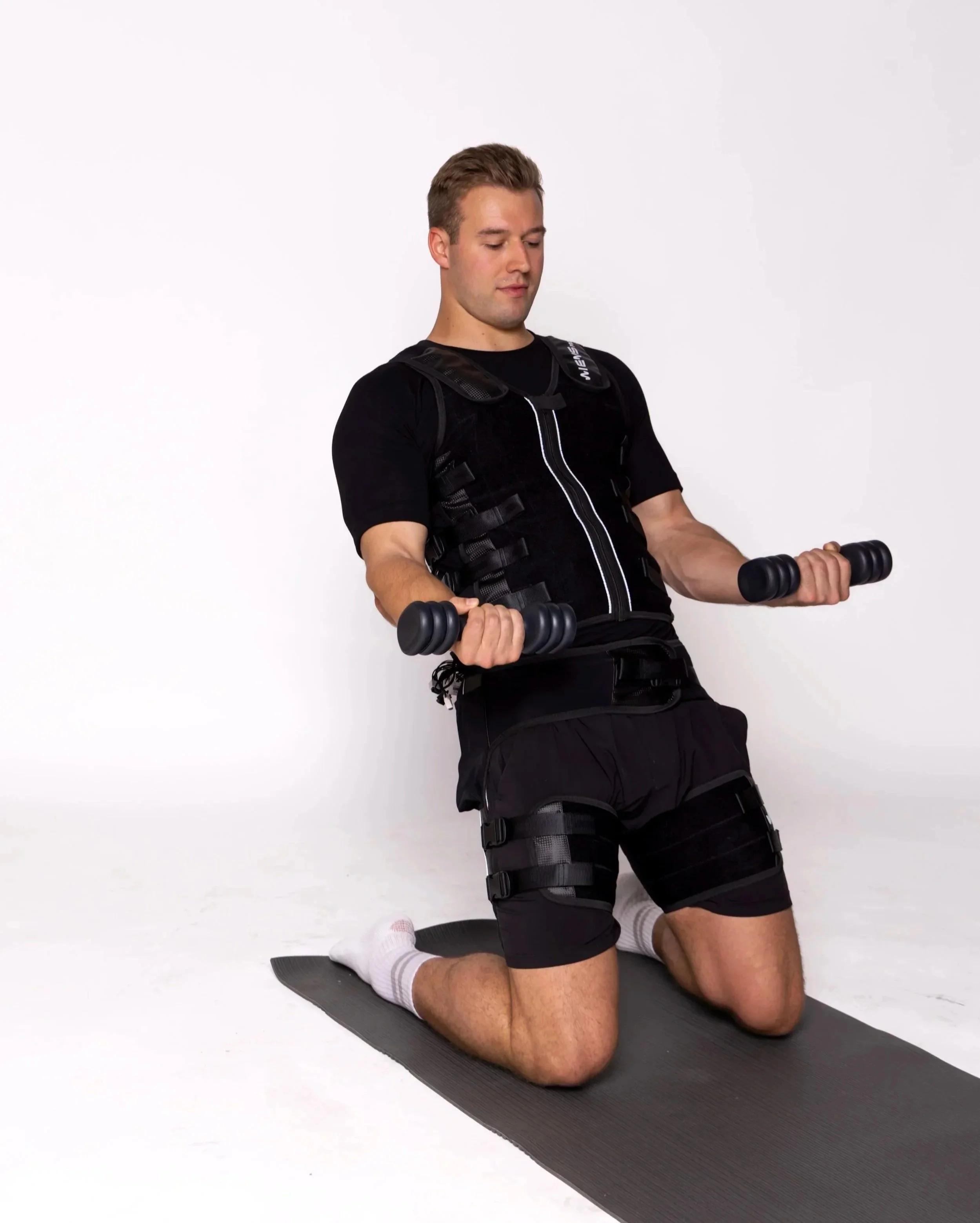 Young man kneeling on a yoga mat, wearing black athletic gear, holding dumbbells in both hands, in a kneeling workout pose against a plain white background.