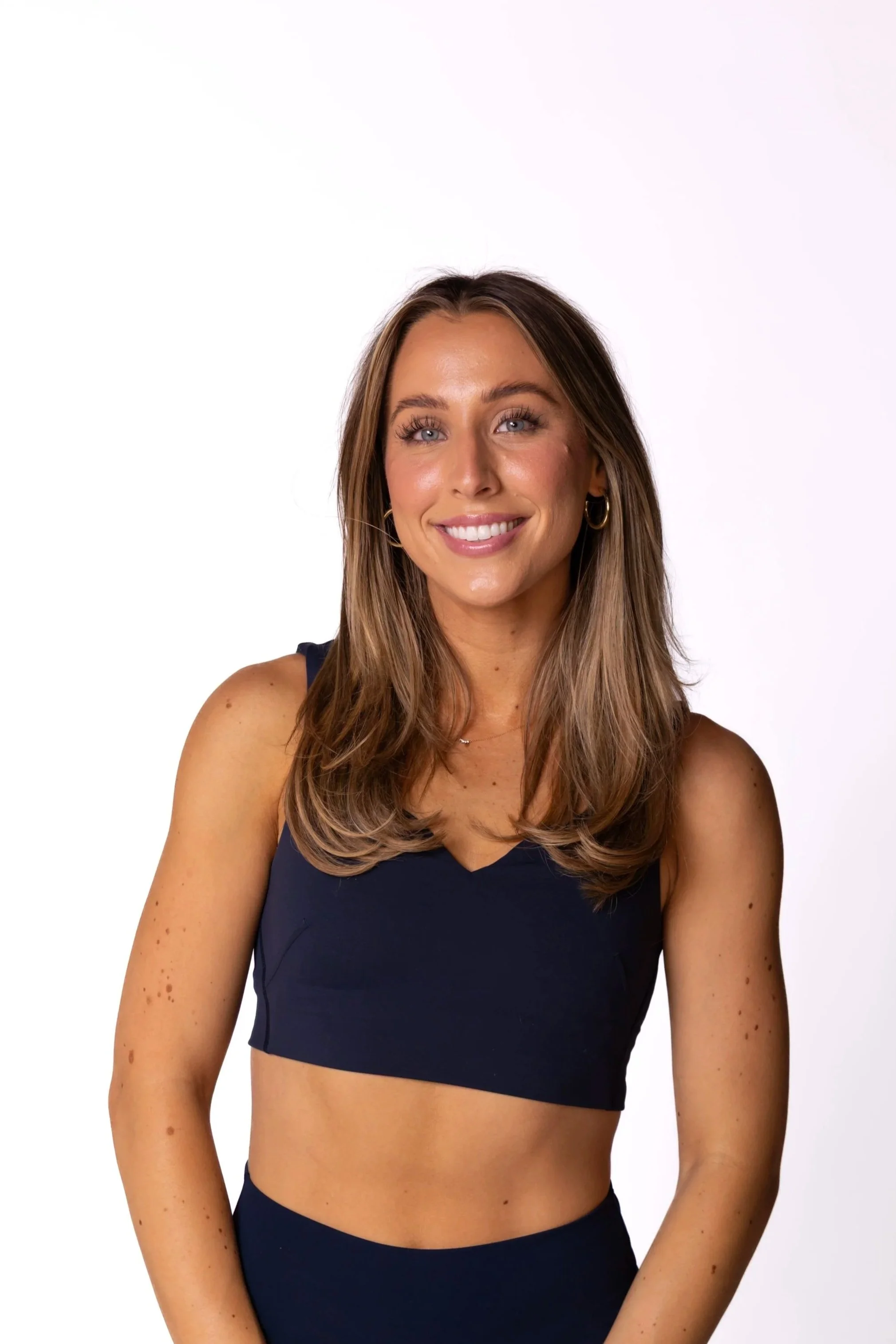 A smiling woman with long blonde hair wearing a white sleeveless crop top and dark pants against a plain white background.