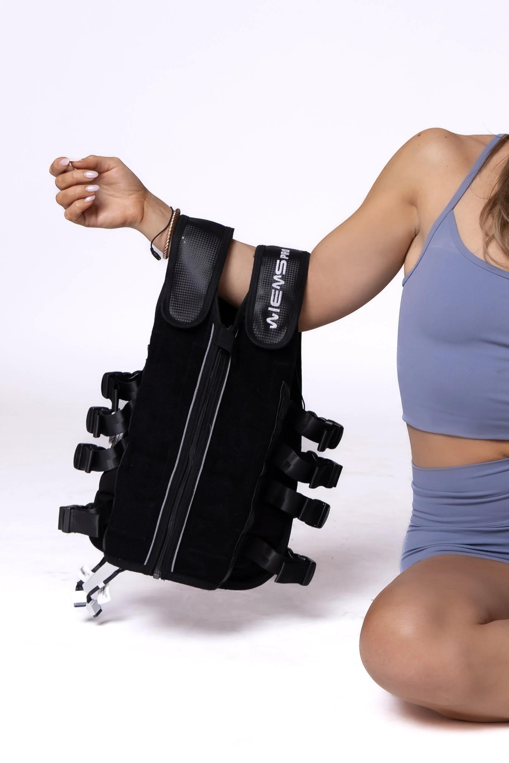 A woman wearing a blue sports bra and shorts, sitting on the floor, holding a black weight vest with multiple adjustable straps and a zipper, against a plain white background.