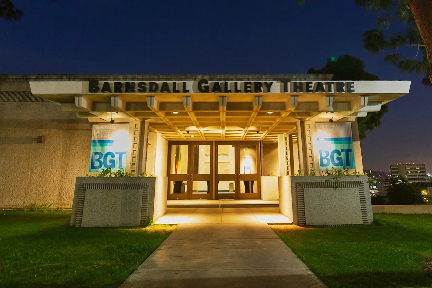 Night view of the entrance to Barnesdall Gallery Theatre, with illuminated glass doors, banners on either side displaying 'BGT,' and a pathway leading to the entrance.
