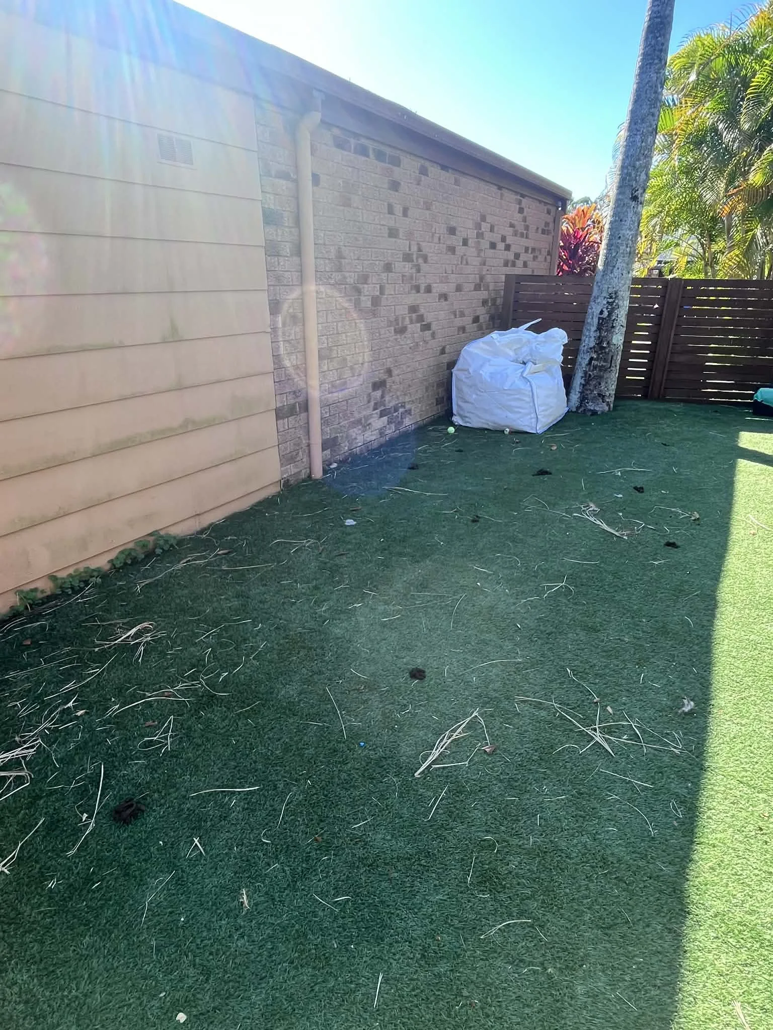 A backyard with artificial grass, a brick wall, a wooden fence, a tree, and a white bag on the ground.
