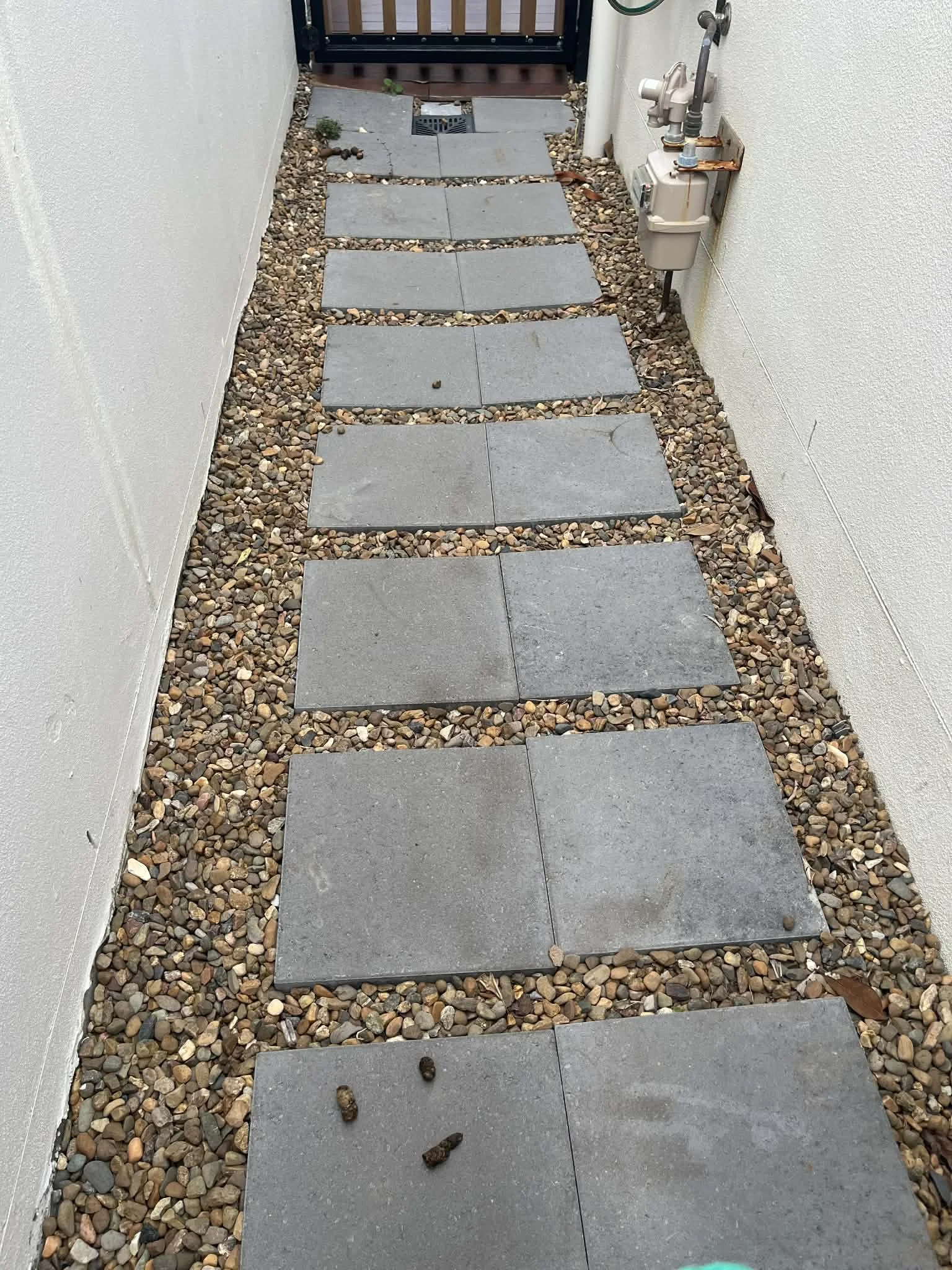 A pathway made of gray square stepping stones surrounded by small pebbles, leading to a gate. A utility meter is mounted on the right wall.
