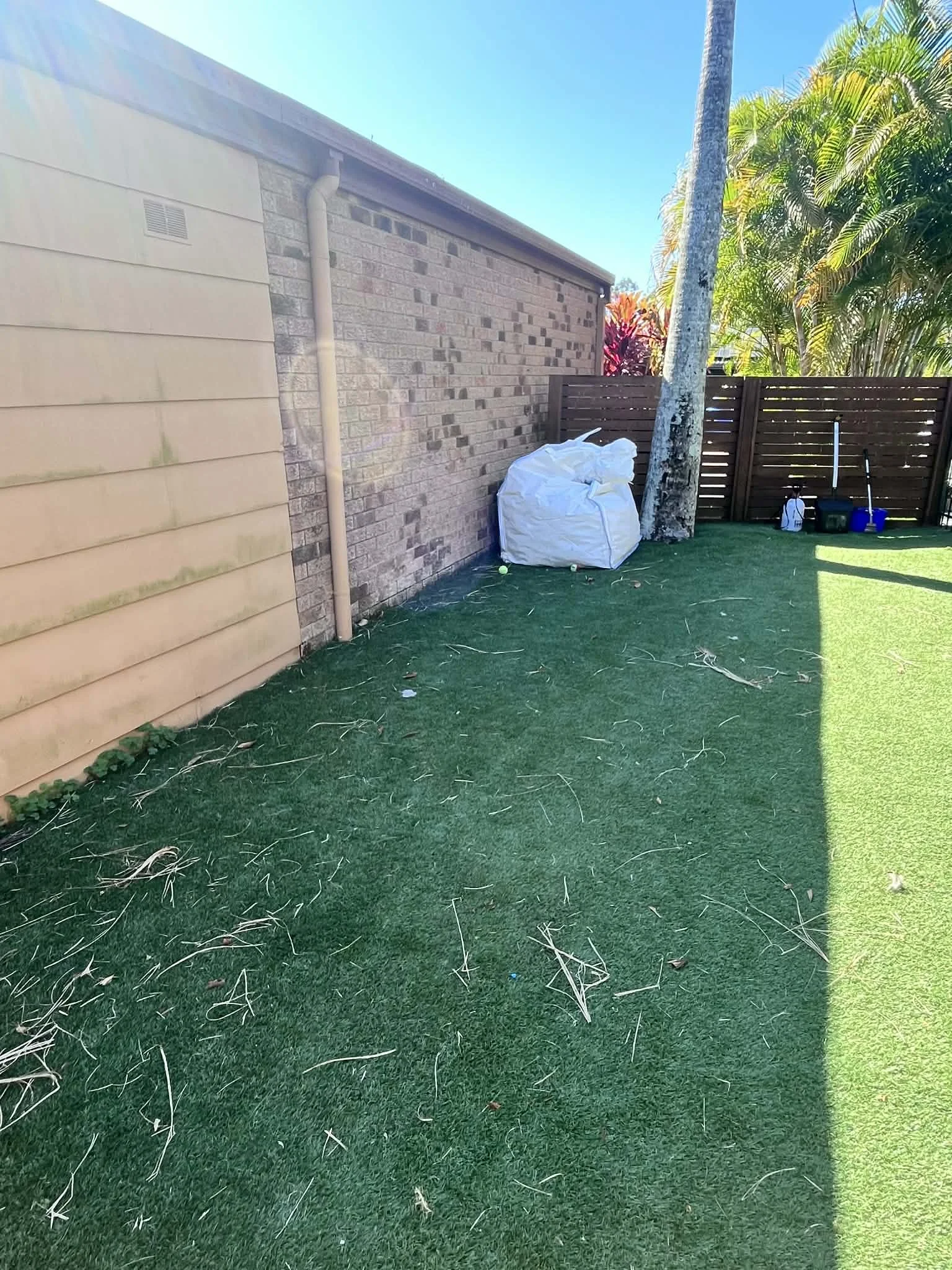 Backyard with artificial grass, a wooden fence, a tree, a white bag, and some tools near the fence.