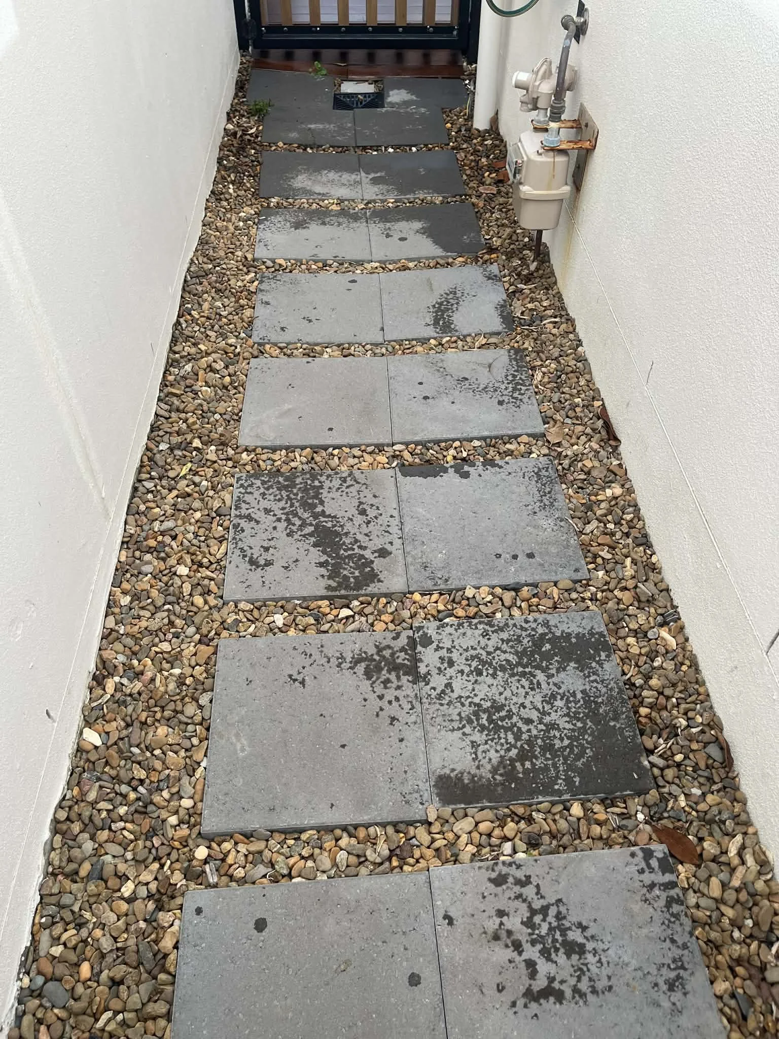 A pathway made of large rectangular gray stones, wet with rain, running between white walls, with small multicolored pebbles filling the gaps around the stones.