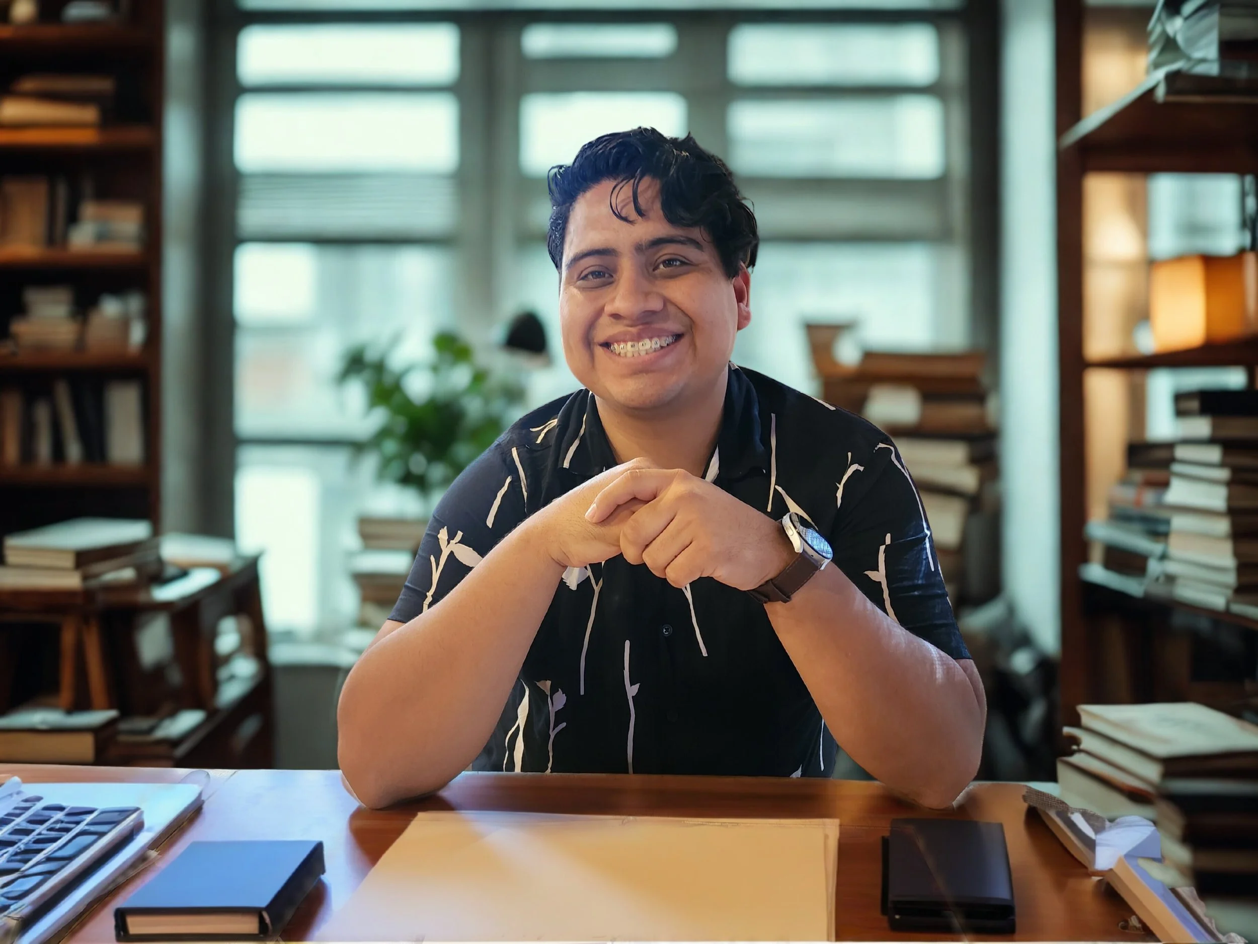 Joven sonriendo sentado en una mesa de oficina rodeado de estantes llenos de libros y papeles.