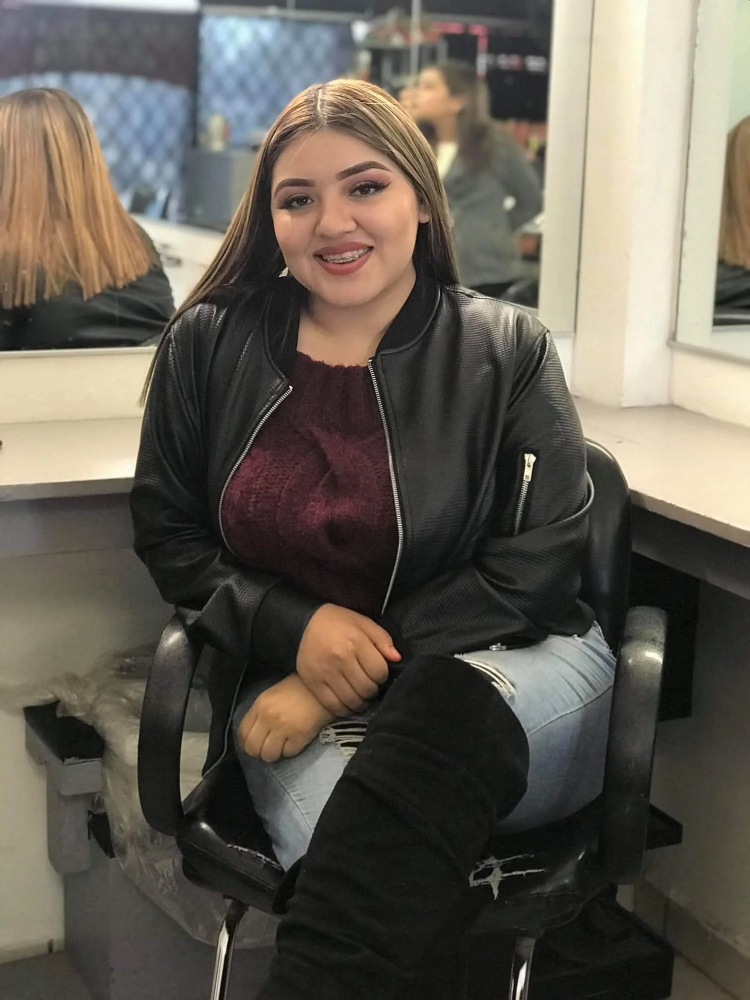 Una joven sentada en una silla frente a un espejo en un salón de belleza o peluquería, con cabello castaño y maquillaje, vestida con una chaqueta de cuero negro, una blusa de color burdeos y pantalones de mezclilla rotos.