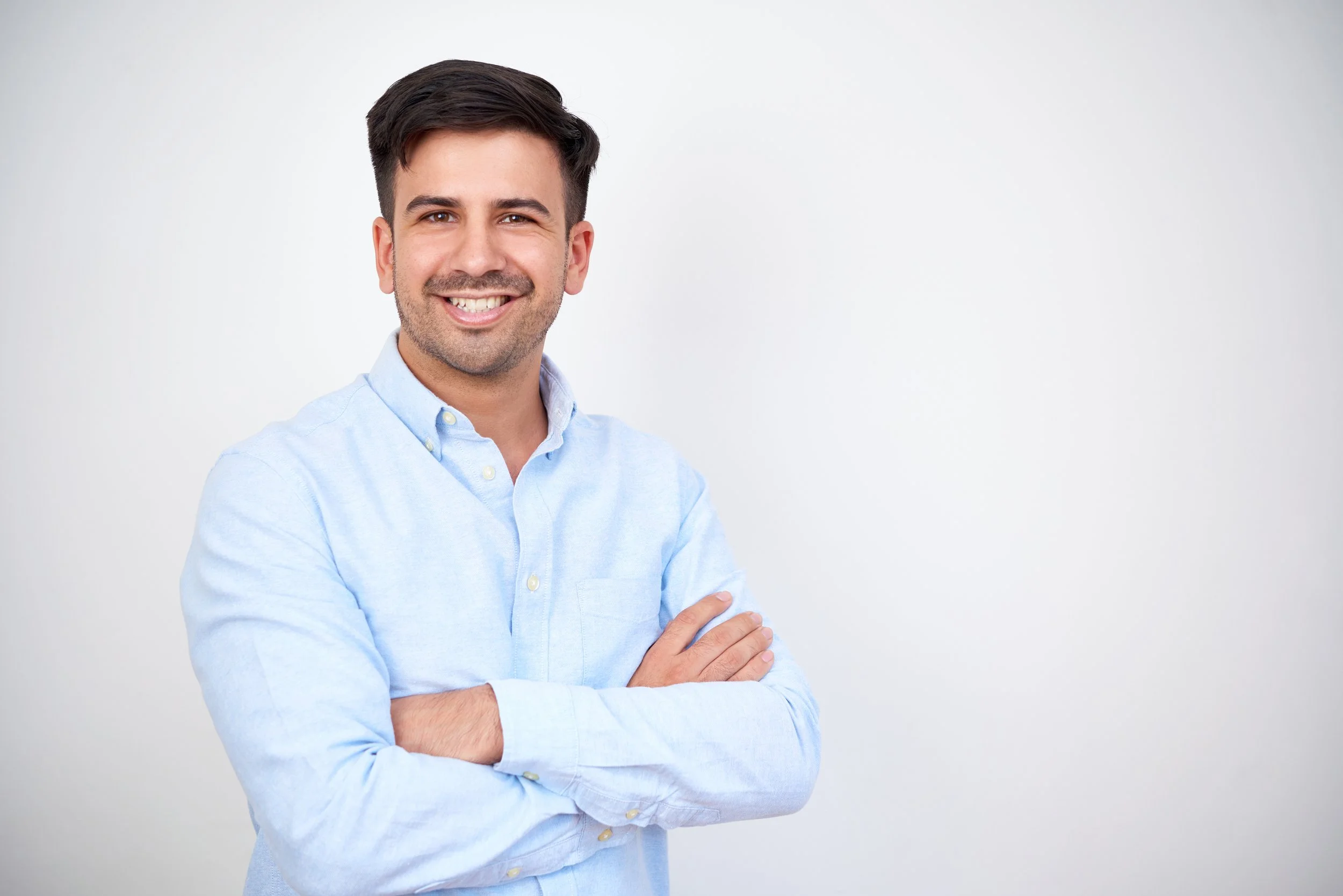 Joven hombre sonriendo, con camiseta de manga larga azul, brazos cruzados, fondo blanco.