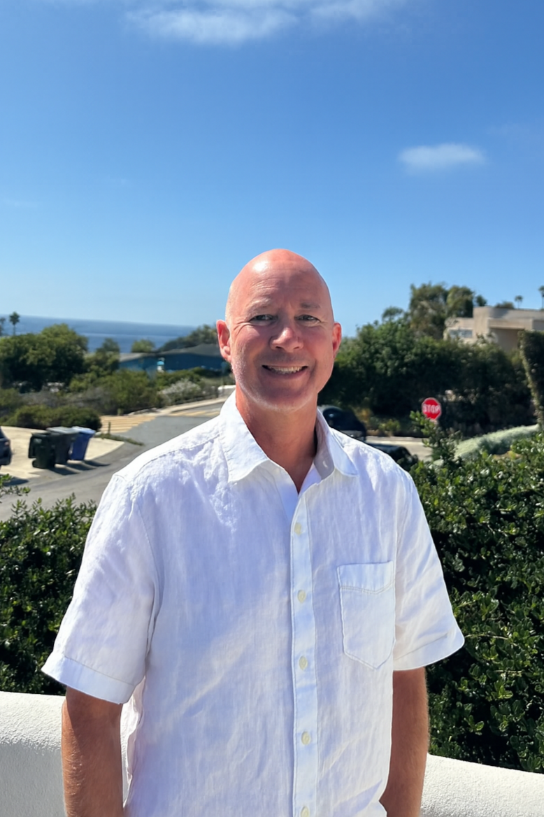 A smiling man in a white shirt standing outdoors on a sunny day with trees, houses, parked cars, and a stop sign in the background.