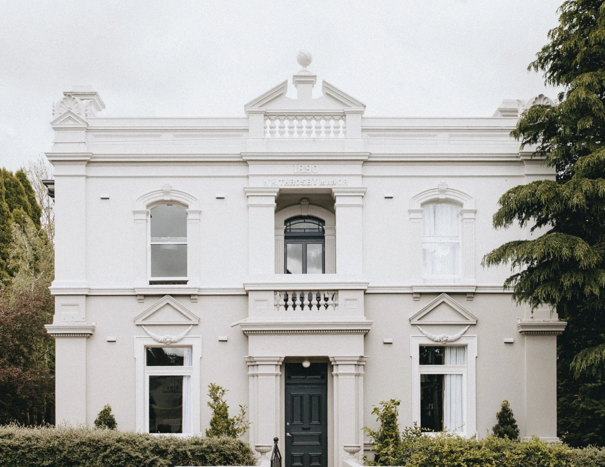 A white, two-story Victorian style house with ornate decorative elements and a black front door, surrounded by green bushes and trees.