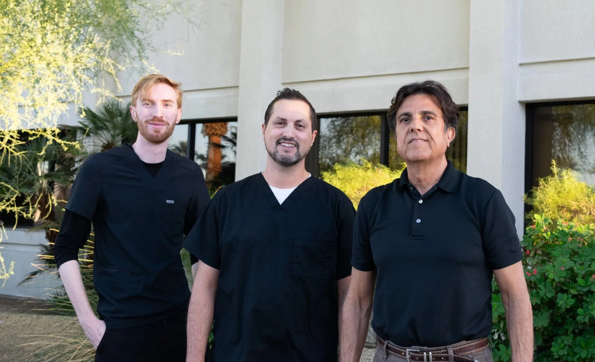 Three male healthcare professionals standing outdoors in front of a modern building with large windows and greenery, wearing black medical scrubs and polo shirts.
