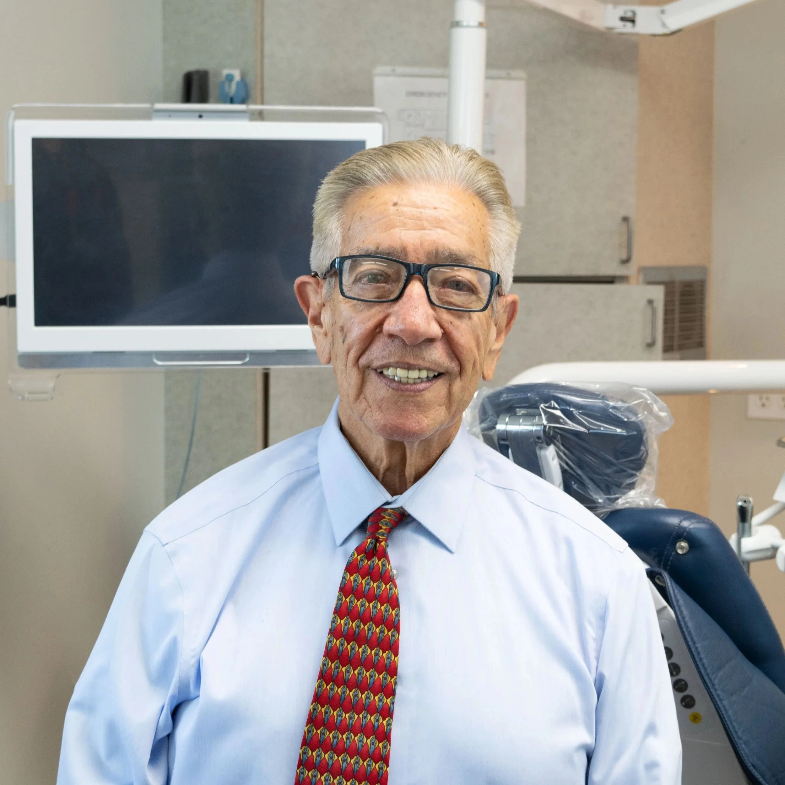 An elderly man with glasses smiling in a dental clinic, wearing a light blue shirt and a colorful tie, with dental equipment and a monitor in the background.