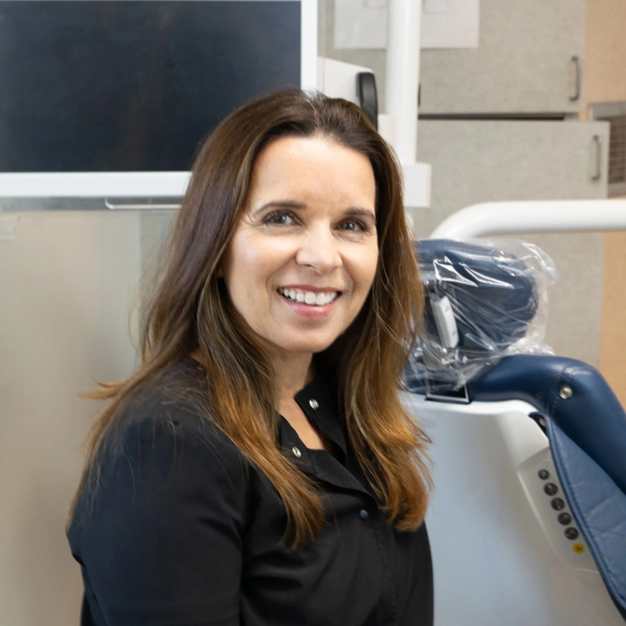 A smiling woman with long brown hair sitting in a medical or dental office, next to a dental chair wrapped in plastic.