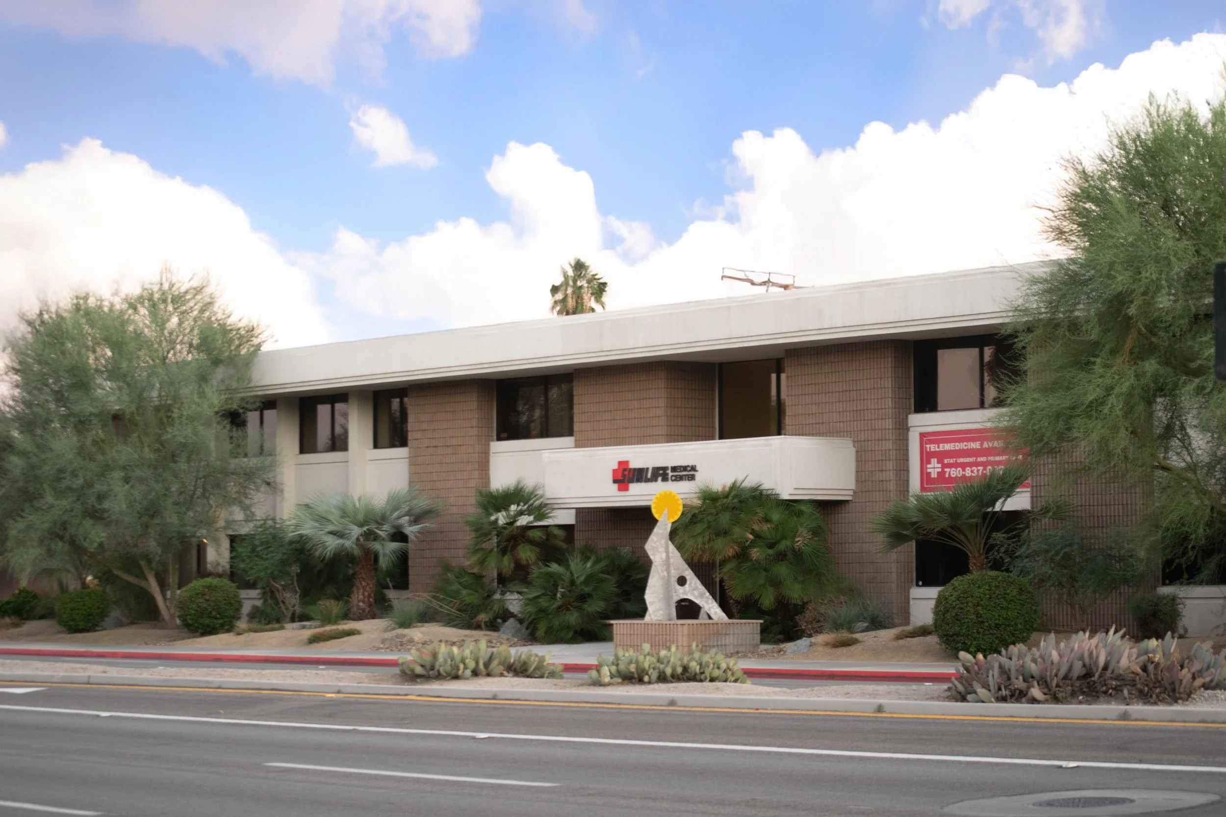 A two-story brick medical building with a white sign that reads 'St. Luke Medical Center.' In front, there are desert plants, including cacti and bushes, and a public art sculpture resembling a giraffe with a yellow circle on top. The sky is partly cloudy.