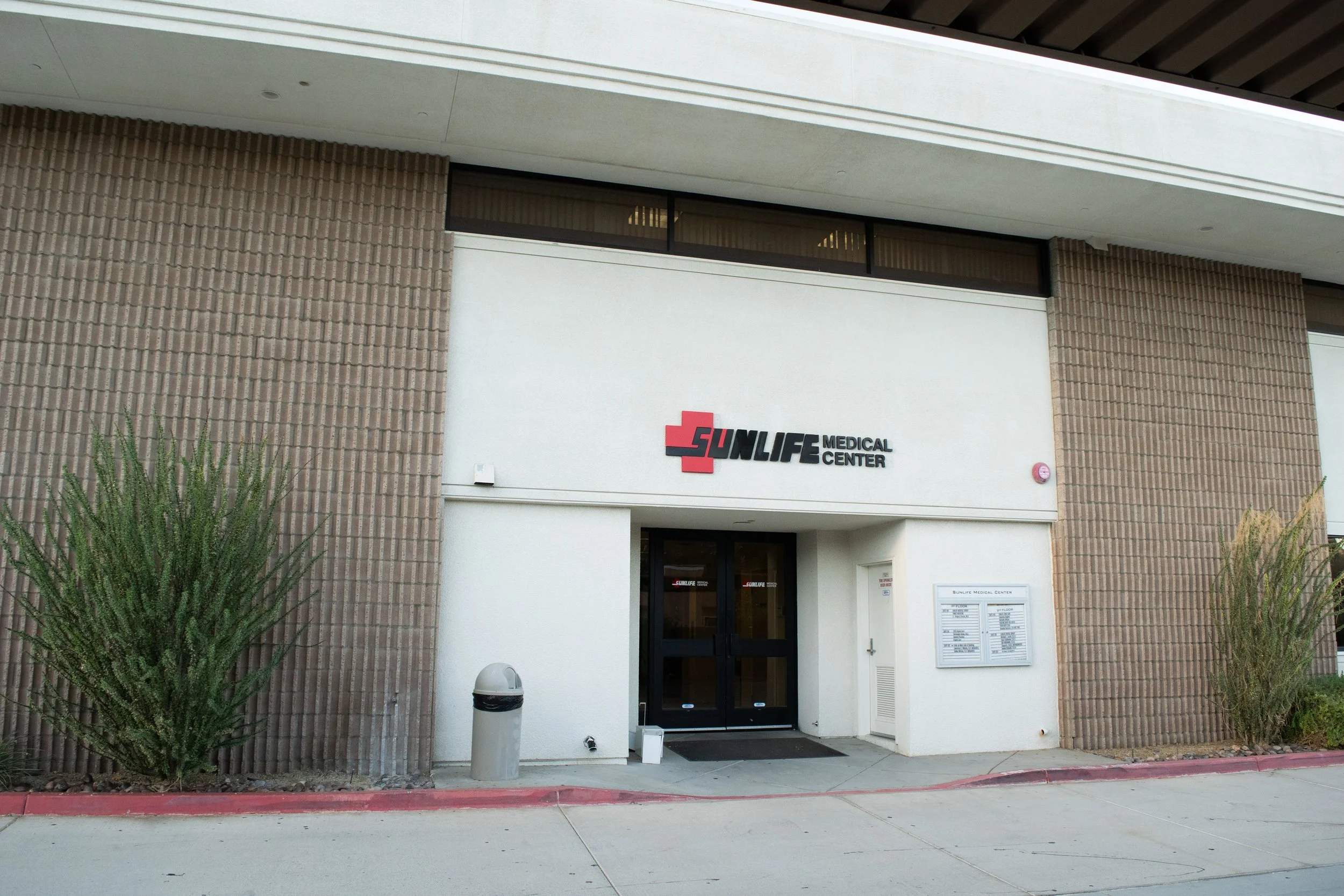 Exterior view of SunLife Medical Center building entrance with sign, plants, trash can, and window.