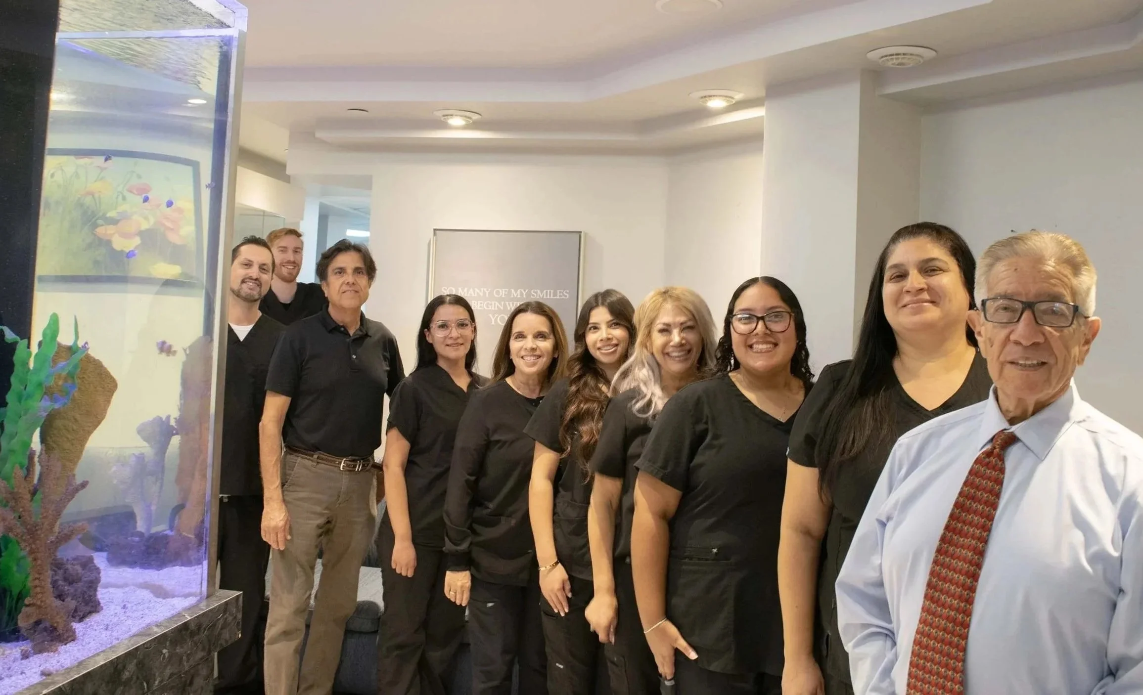 Group of eight staff members with diverse appearances, smiling and standing in a line indoors near an aquarium, with a wall sign that reads 'So many of my smiles begin with you.'