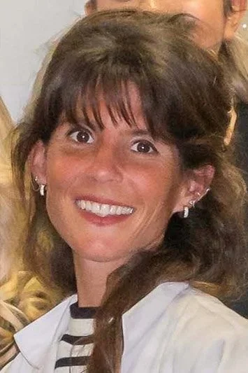 Smiling woman with brown hair and bangs, wearing earrings and a white top with black and white striped collar.