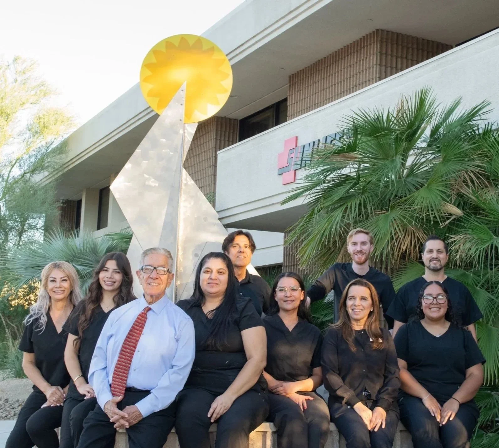 Group of eight healthcare professionals, including doctors and nurses, sitting and standing outside a building with a modern artwork sculpture, palm trees, and a pharmacy sign.