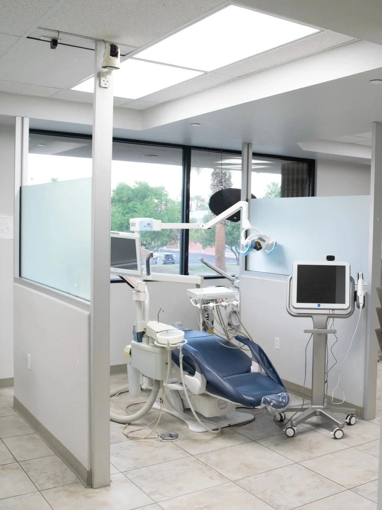 Dental treatment room with dental chair, monitors, and dental equipment, separated by glass partitions, with large windows and trees outside.