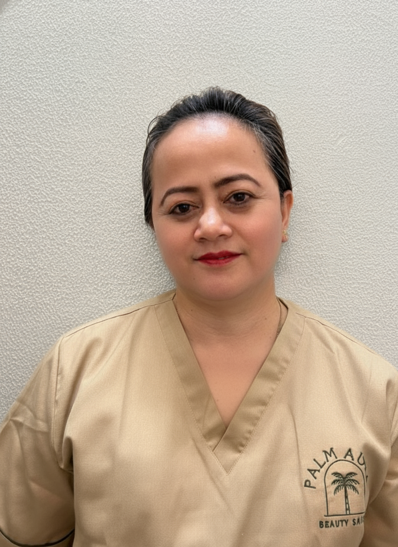 A woman wearing a beige uniform with the Palmau Beauty Spa logo, standing against a textured off-white wall.