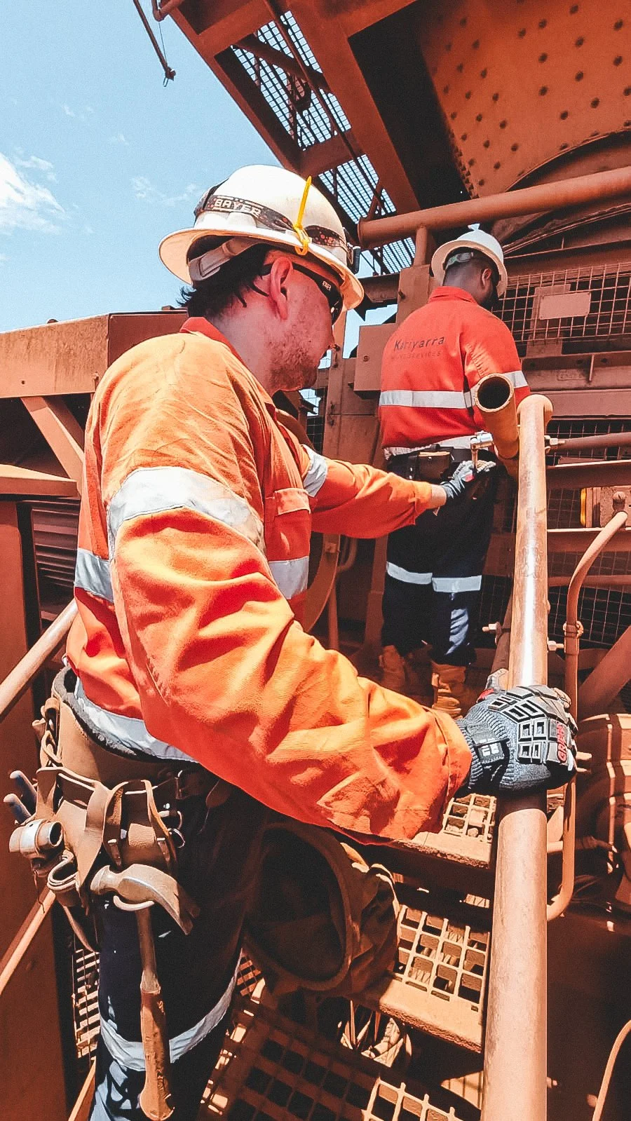Two workers in orange safety gear and white helmets working on a large industrial machine outdoors.