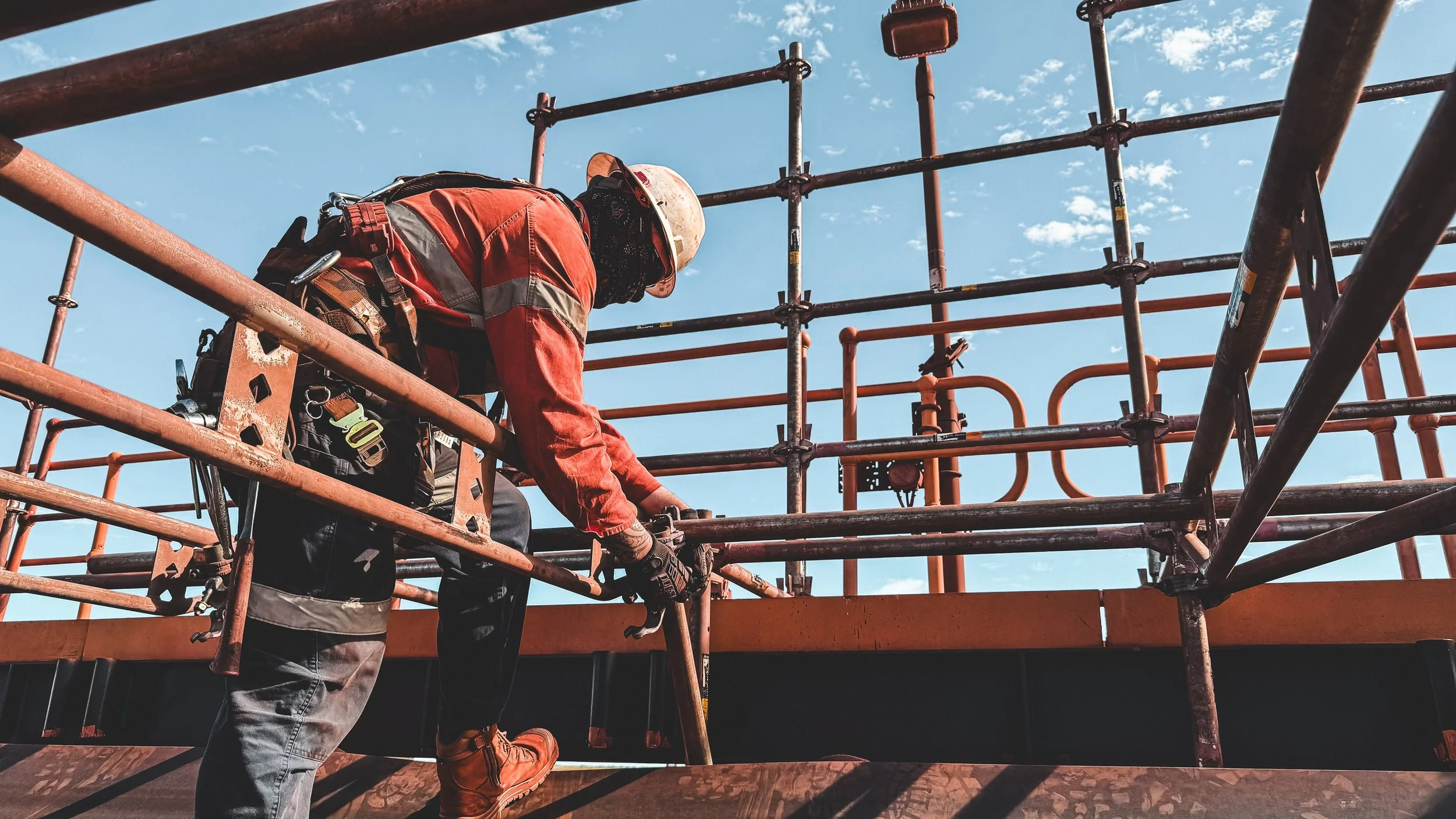 Construction worker climbing orange scaffolding against a blue sky.