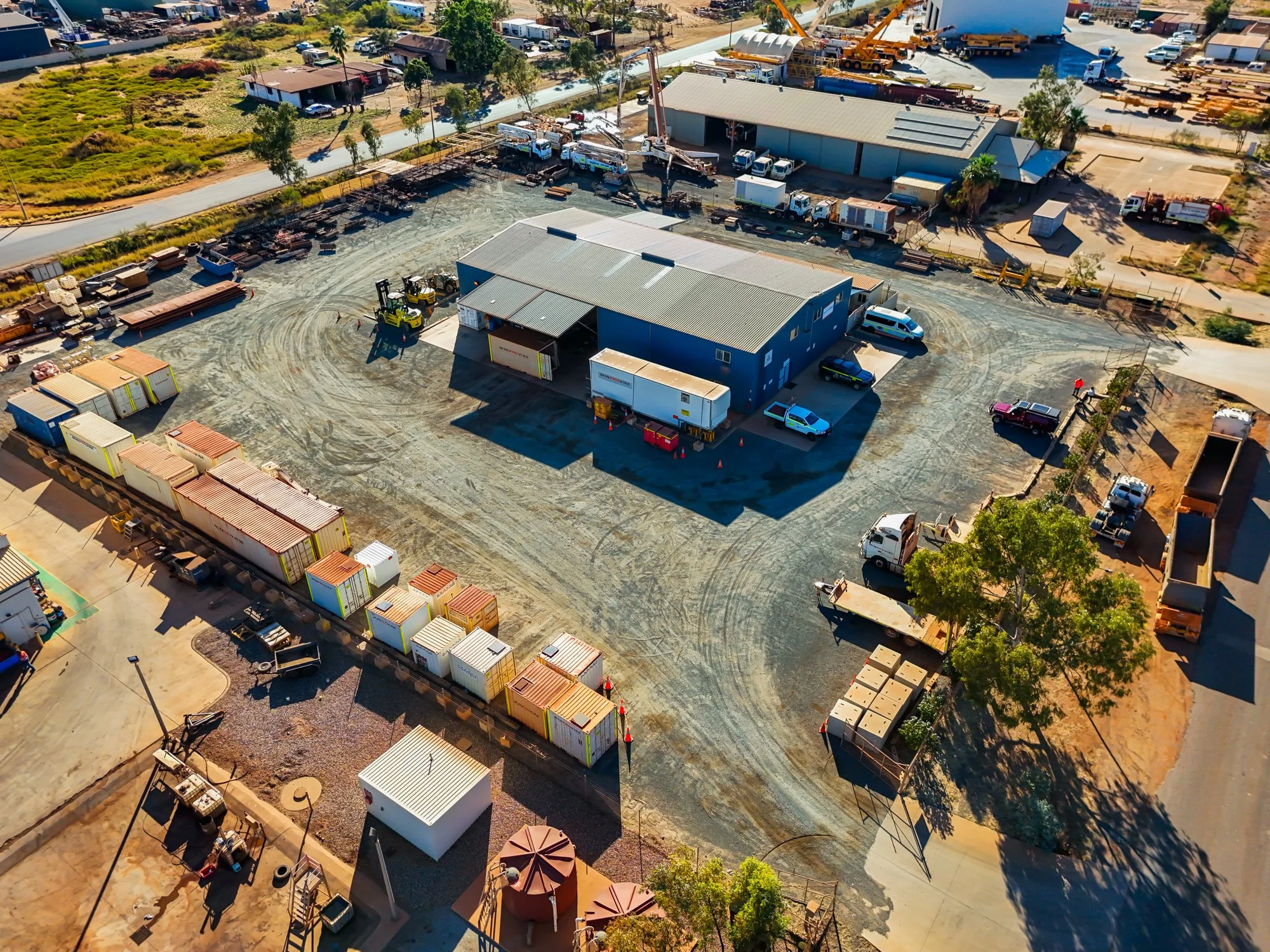 Aerial view of a construction site with a blue building, construction vehicles, and storage containers, surrounded by other industrial buildings and parked trucks.