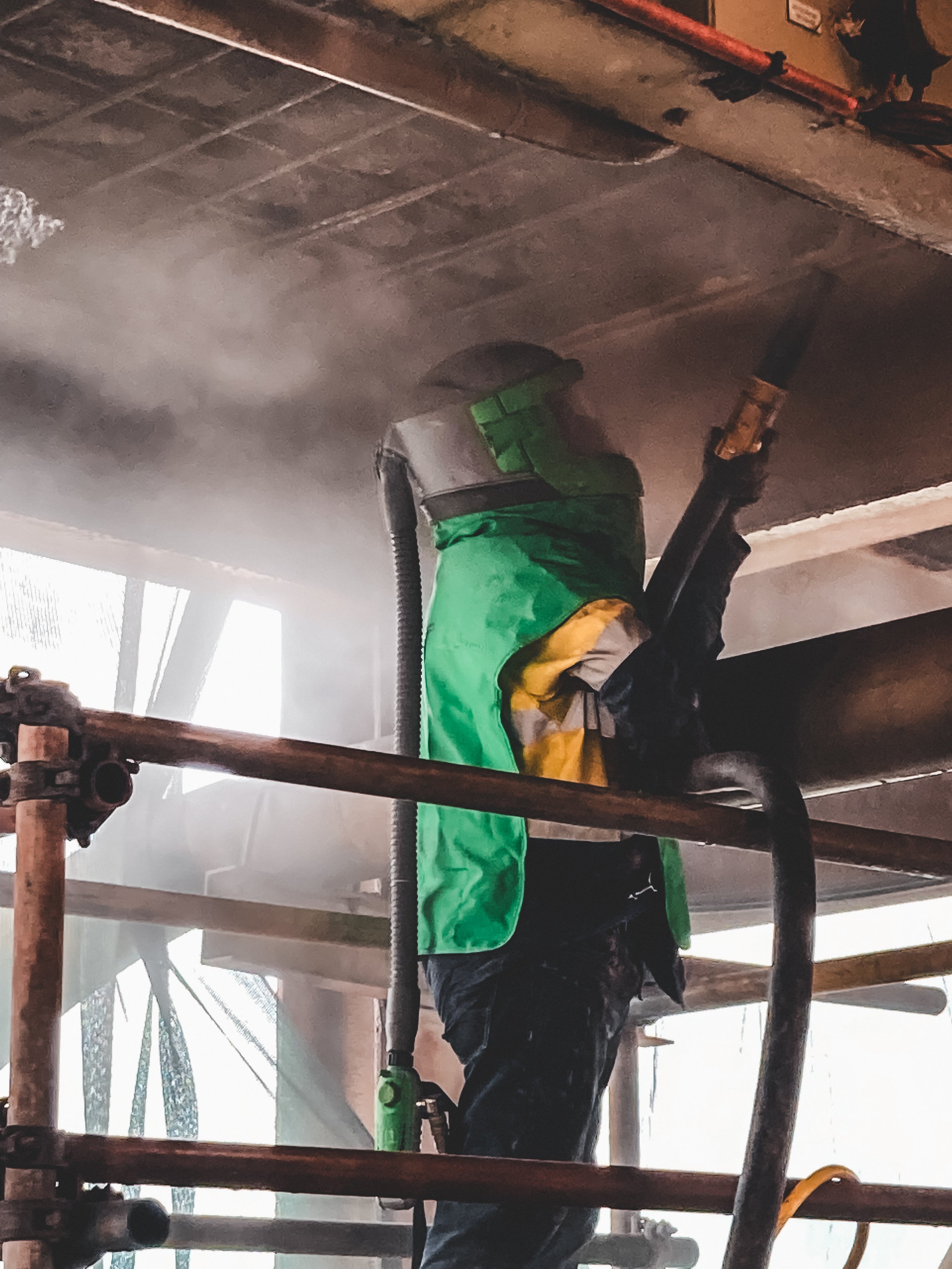Construction worker wearing a helmet and safety vest using a power tool on a ceiling, with dust and smoke surrounding him