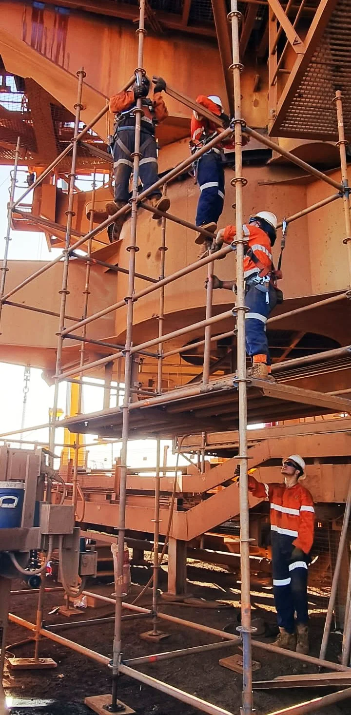 Three construction workers wearing orange and blue safety gear and helmets working on scaffolding at a construction site of a large building or industrial structure.