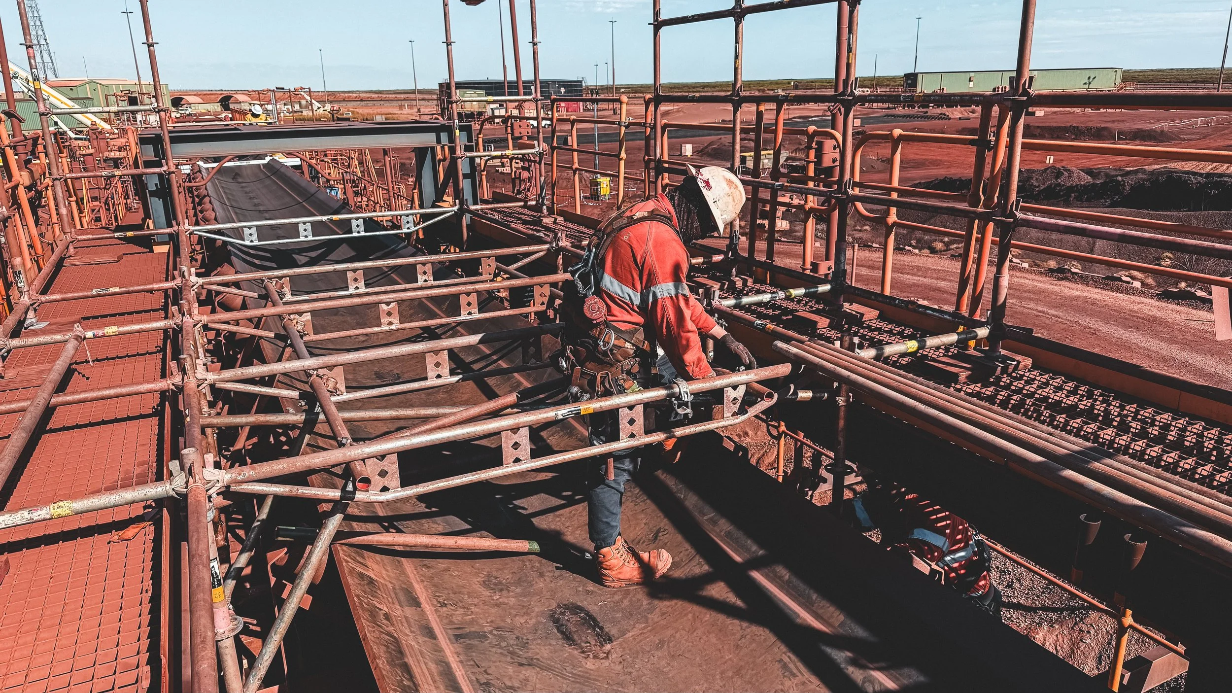 A worker in protective gear working on a large metal structure in an industrial site, possibly a mine or construction area, with a dirt ground and open sky in the background.