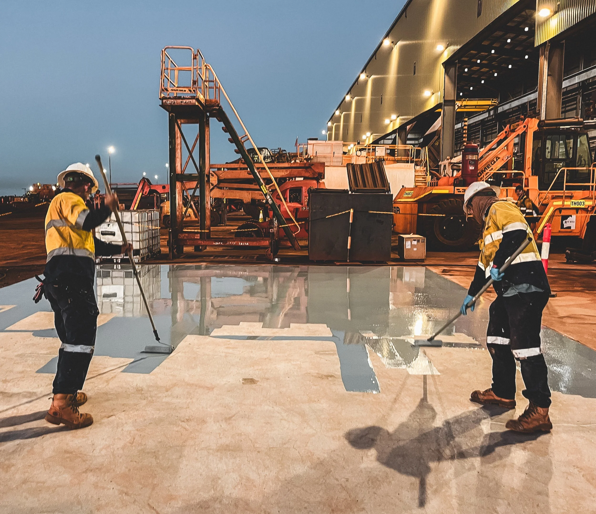 Workers in safety gear applying sealant to a concrete surface at an airport tarmac during evening hours.