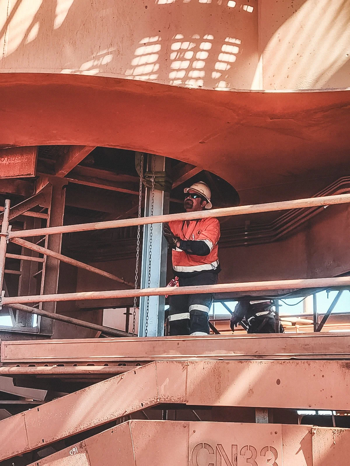 A construction worker wearing safety gear, including a white helmet, sunglasses, and a reflective orange jacket, standing on a raised platform at an industrial site.