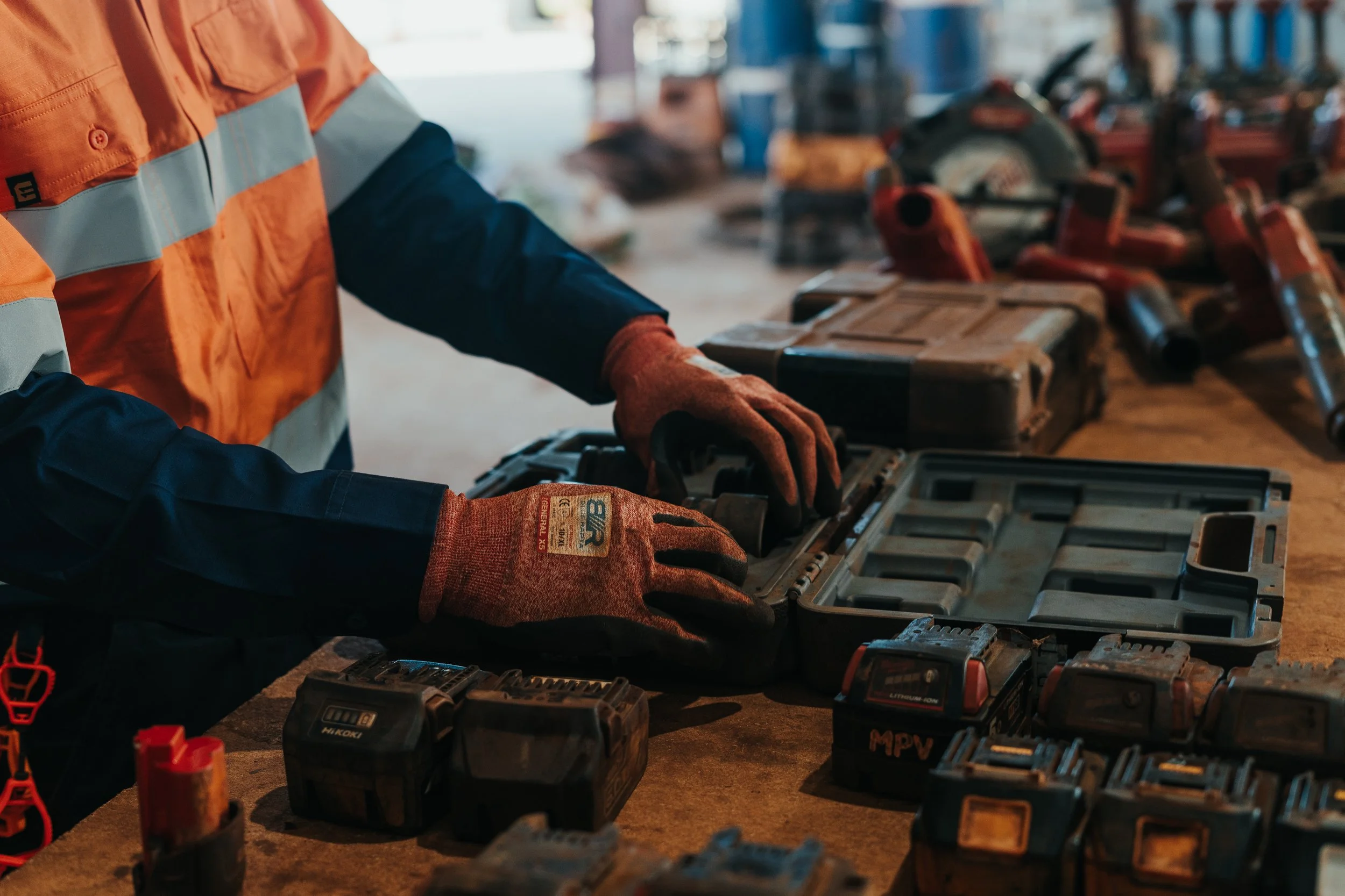 Person wearing safety gloves and a reflective safety jacket working on a large toolbox filled with power tools and batteries in a workshop.