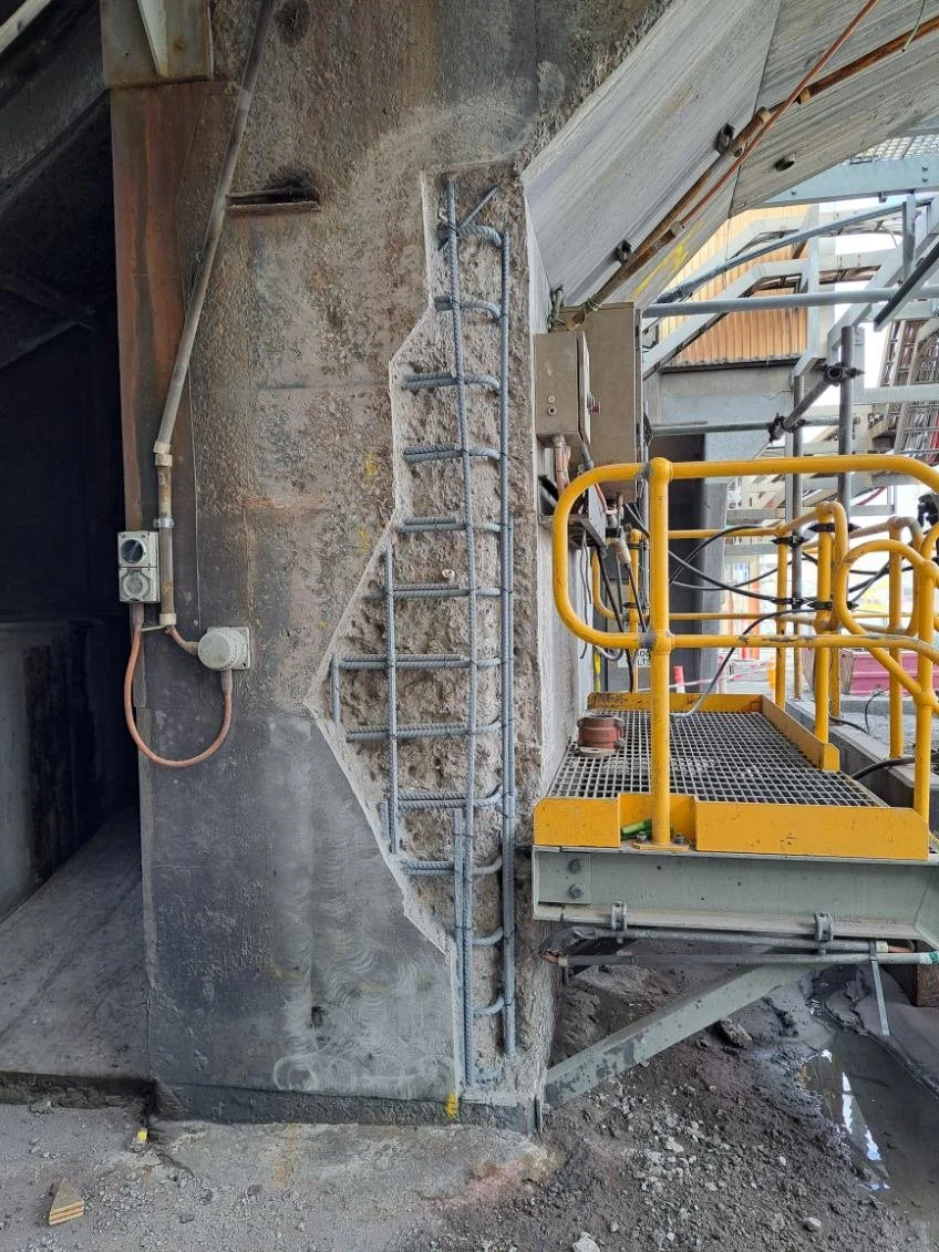 Construction site showing a concrete wall with a metal ladder attached, electrical box and wiring, and a yellow lift platform on a scaffolding structure.