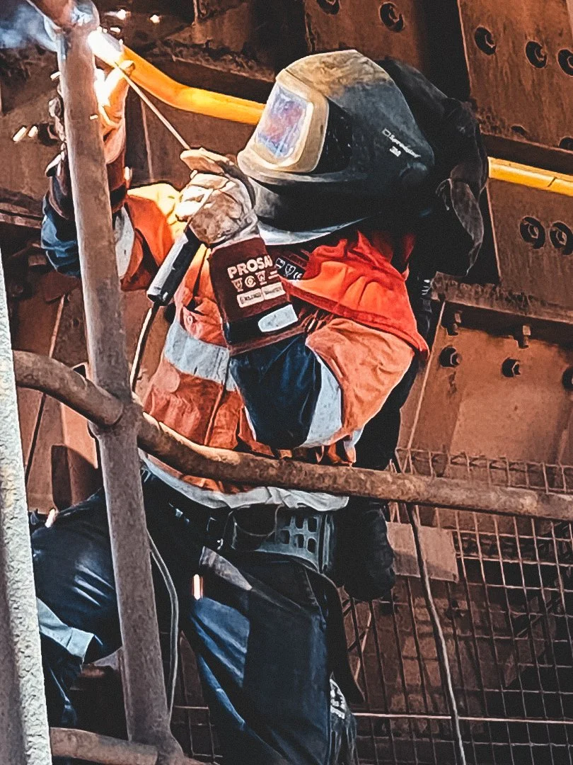 A worker welding metal on an industrial construction site, wearing a protective welding helmet, gloves, and safety vest.