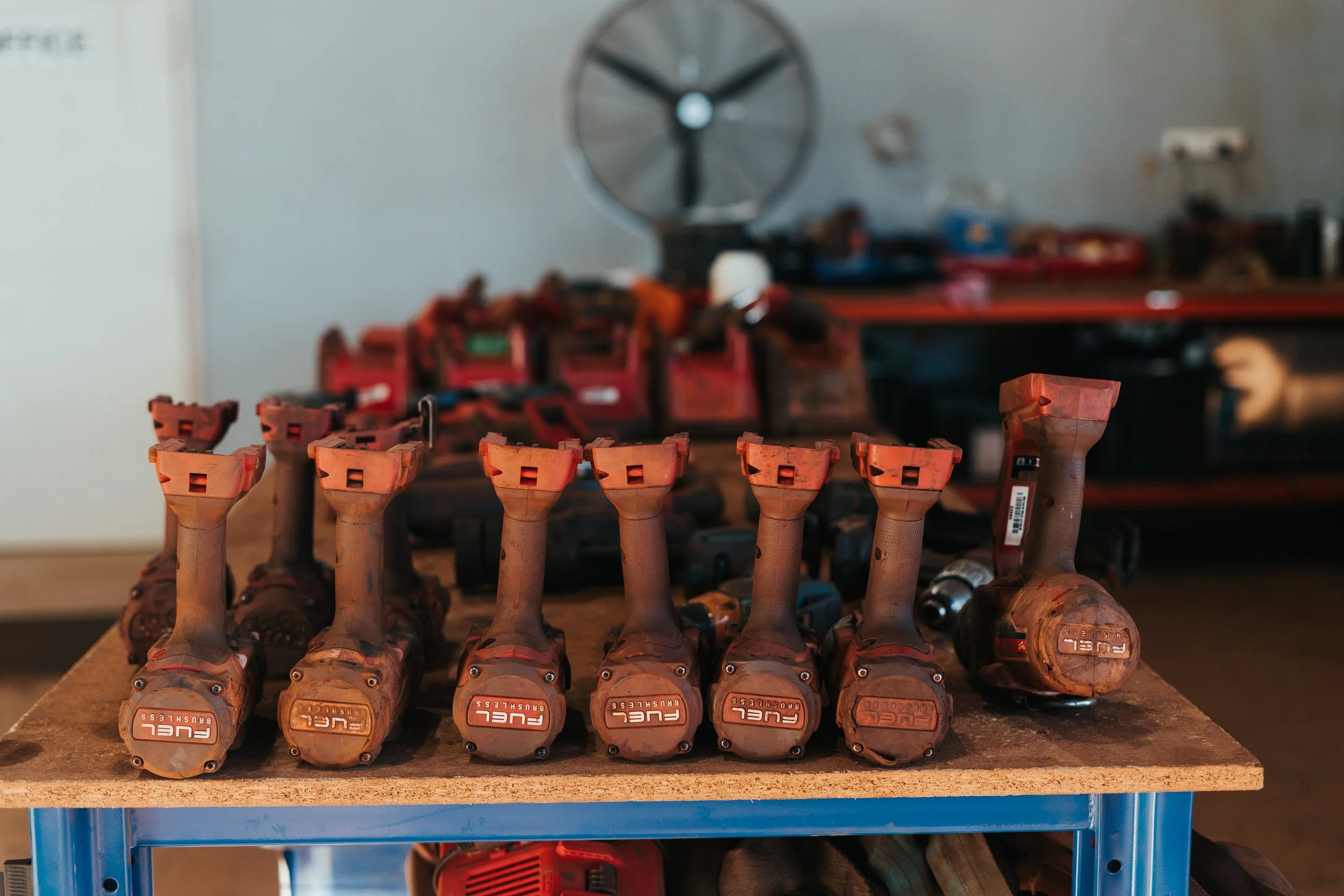 Seven used power tool battery packs lined up on a work table in a workshop, with additional tools and equipment in the background.
