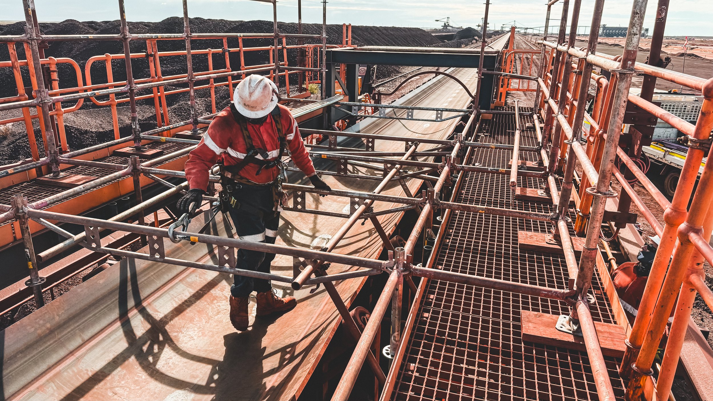 A construction worker wearing a safety helmet and harness working on scaffolding at an industrial site with open ground and soil piles in the background.