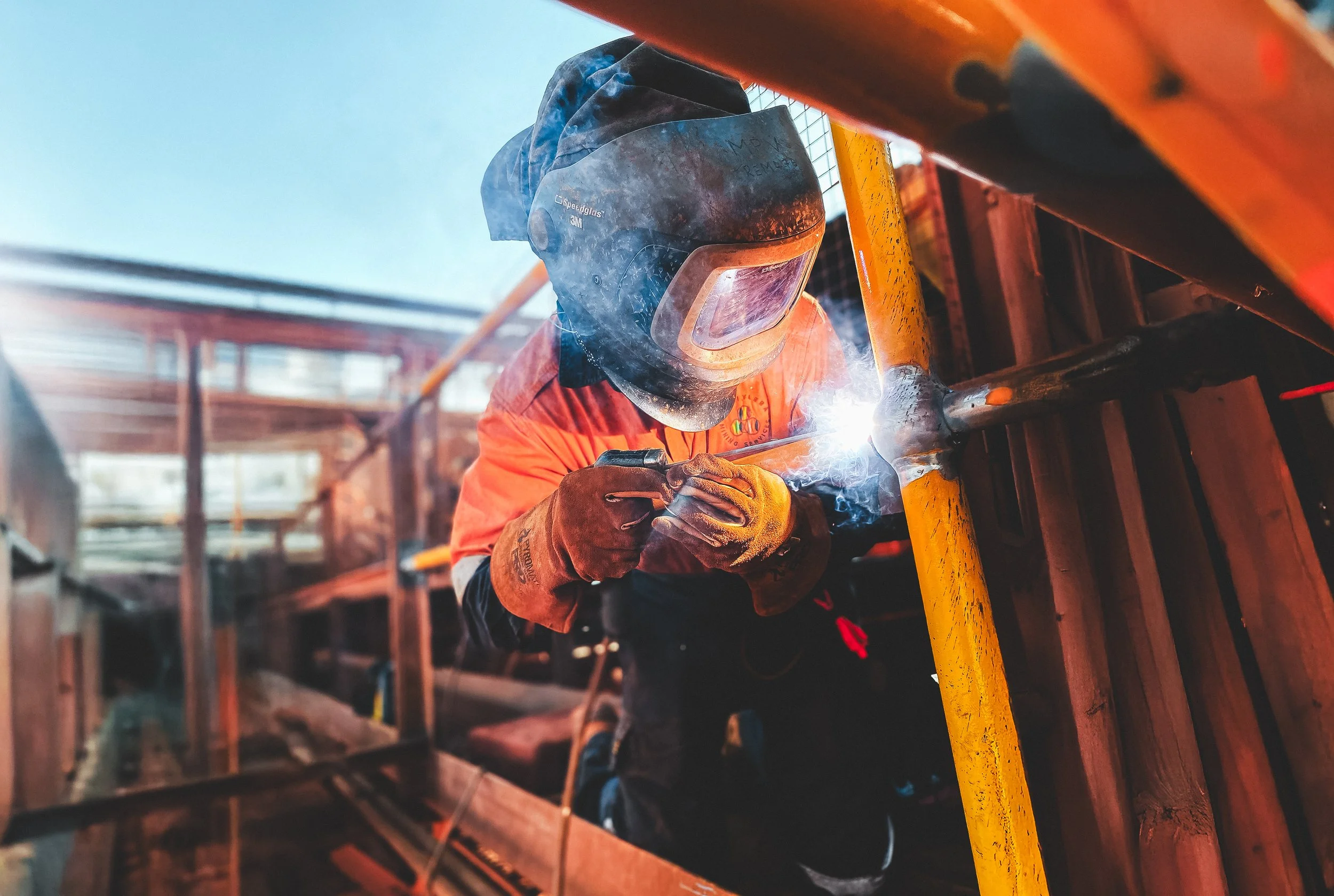 A construction worker wearing a welding helmet and orange safety jacket welding a metal pipe at a construction site.