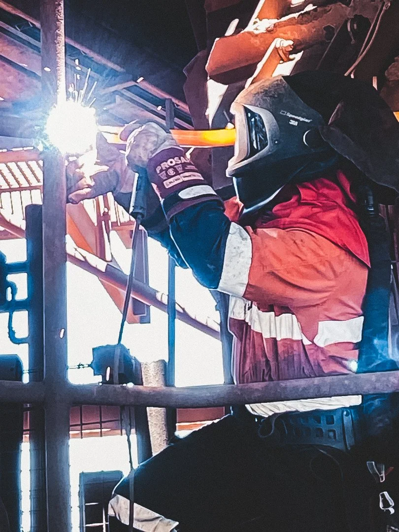 A person in red and white welding gear is welding metal on a structure, with bright sparks flying from the welding torch and the person wearing a welding helmet.