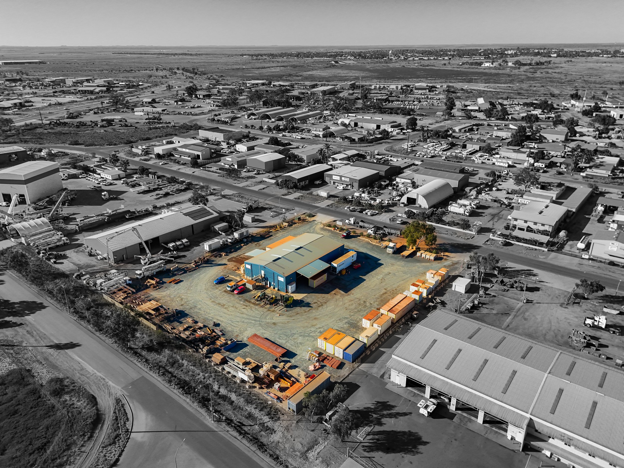 Aerial view of an industrial area with a color-highlighted building and storage containers, surrounded by a parking lot and other buildings, with a road in the foreground and a large open space in the background.