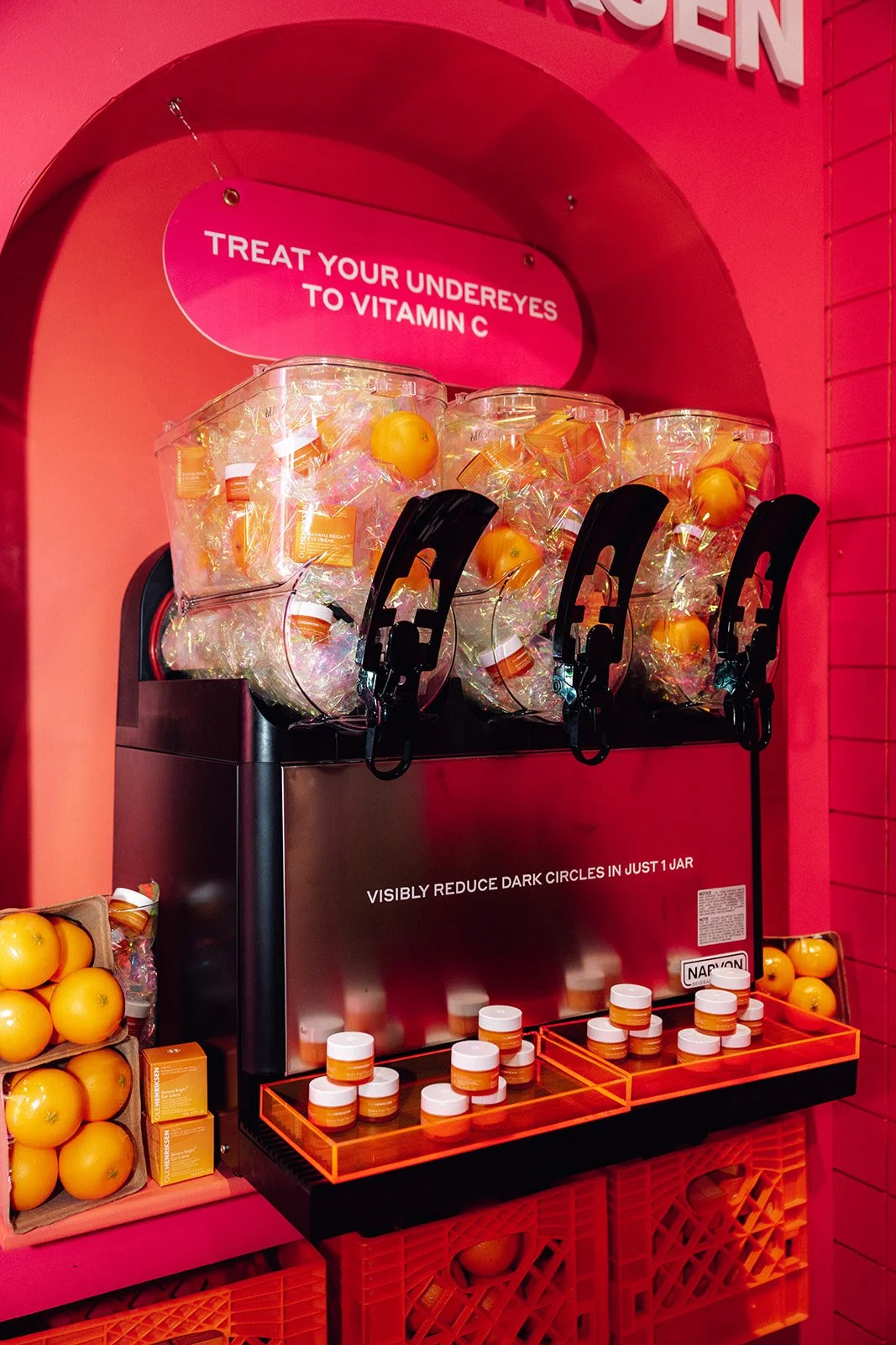 A display of vitamin C supplements in a store with a sign encouraging to treat under eyes to vitamin C. The display has containers of orange-colored tablets or capsules, with some on shelves and others in clear plastic containers, set against a pink 